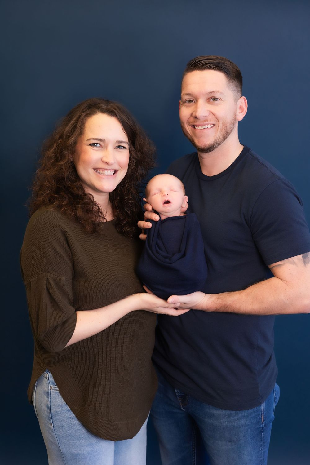 A proud couple holds their newborn baby wrapped in navy blue against a dark backdrop during a studio photoshoot.