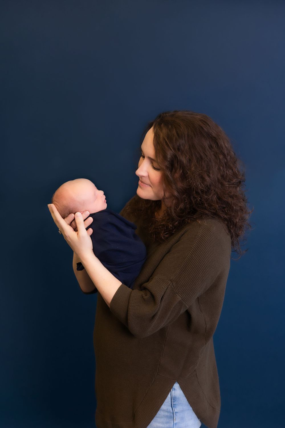 A woman in a brown sweater holds a newborn baby wrapped in navy blue against a dark blue background.