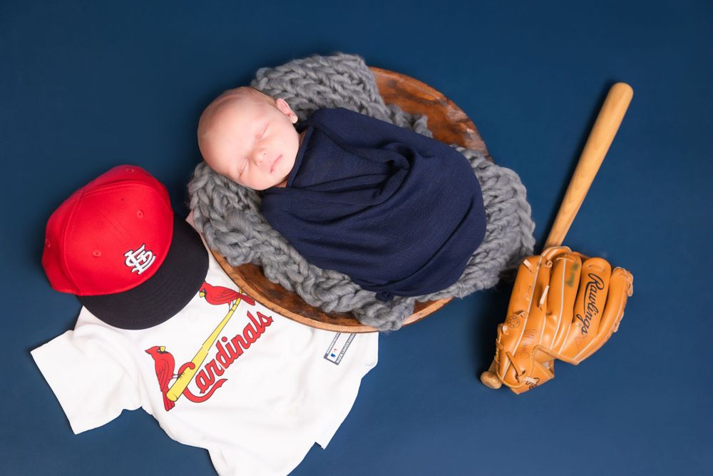 Baby wrapped in a blue blanket with St. Louis Cardinals baseball gear including a jersey, cap and glove nearby.
