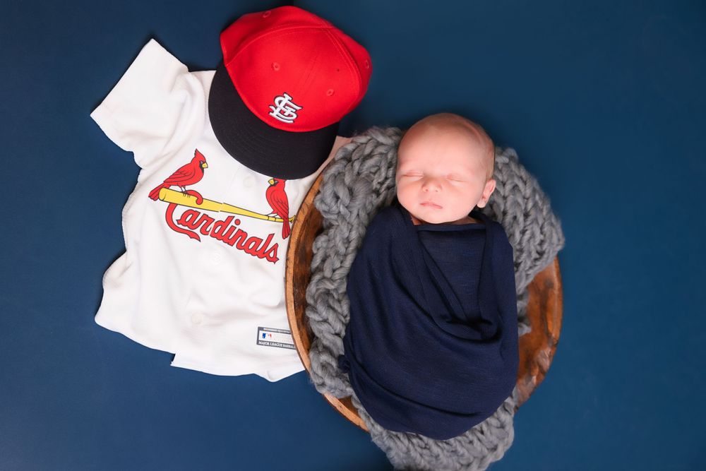 Baseball-themed baby photo featuring a red Cardinals cap, white jersey and infant in navy wrap on blue background.