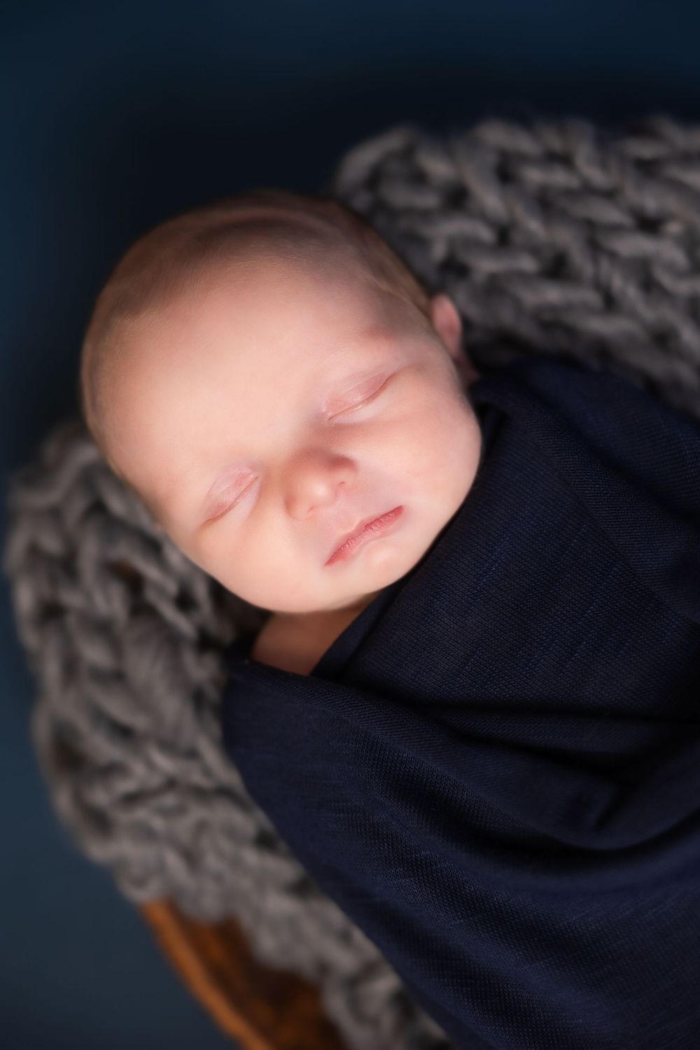 A sleeping newborn baby swaddled in navy blue fabric rests peacefully on a chunky knit gray blanket.