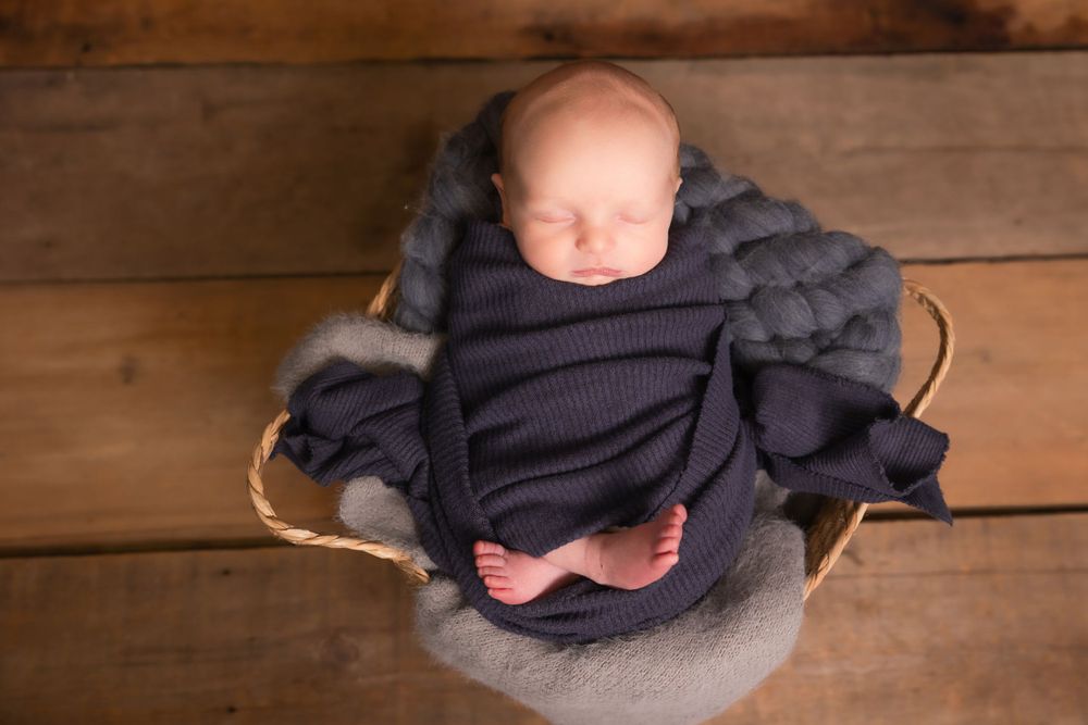 Sleeping newborn baby wrapped in navy sweater lying on grey blanket in wooden basket on rustic floor.