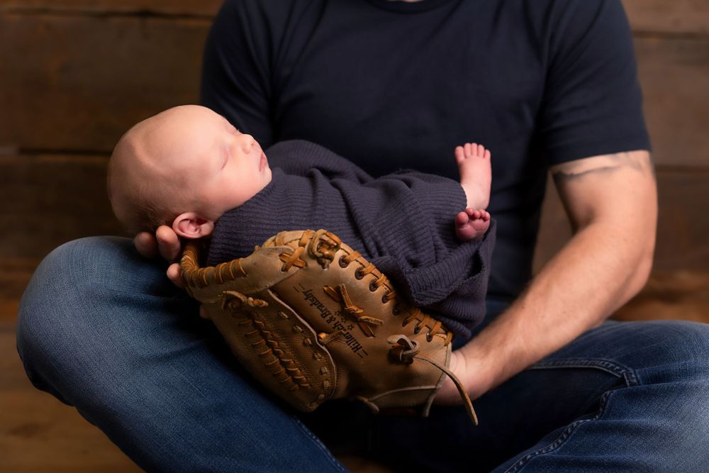 A baby rests peacefully on a baseball glove while lying across someone's lap in casual attire.