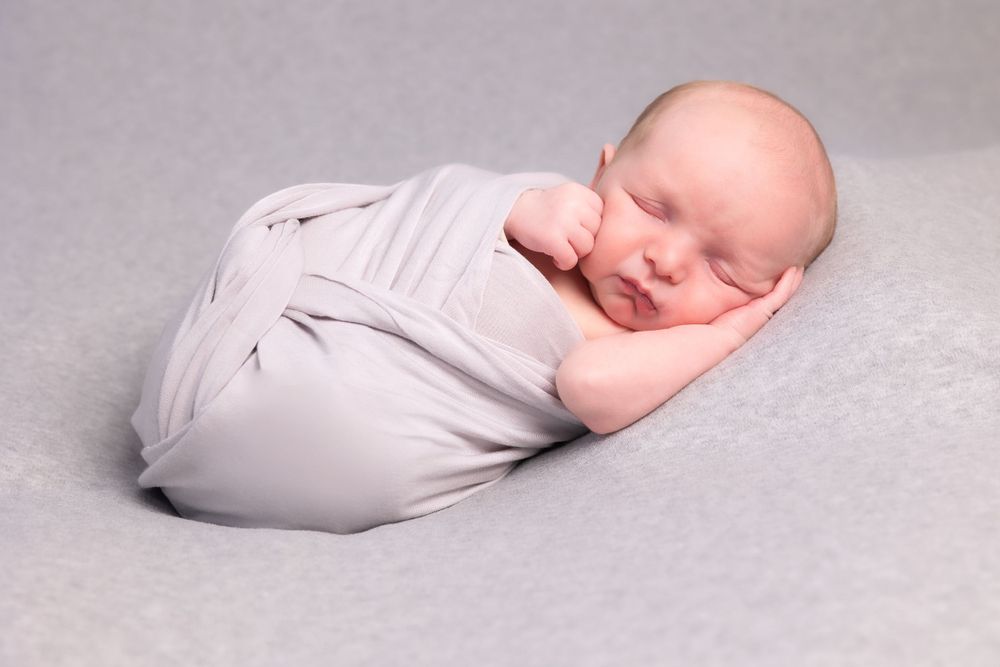 A peaceful newborn baby wrapped in soft white fabric lies sleeping on a gray backdrop.