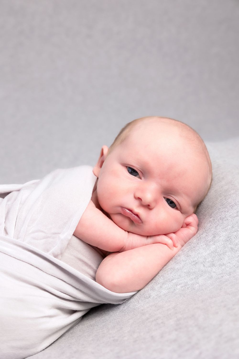 Sweet baby in white outfit rests sleepily with head propped on hands against gray background.