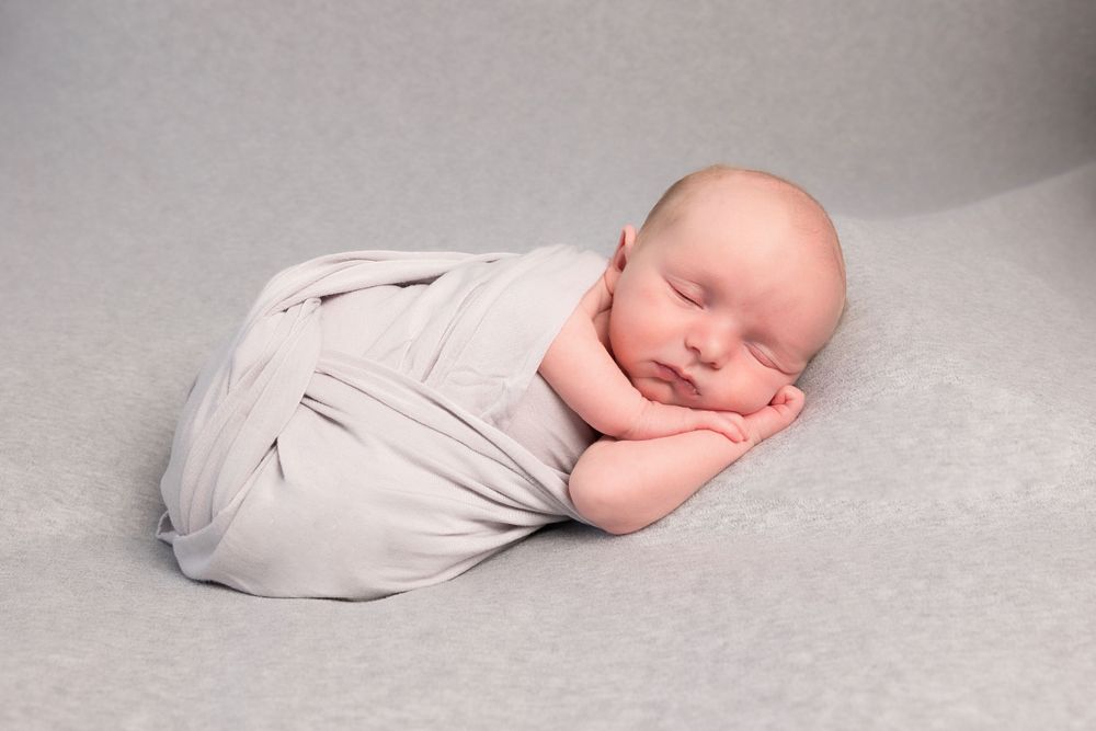 Newborn baby sleeping peacefully while swaddled in soft white fabric on grey background.