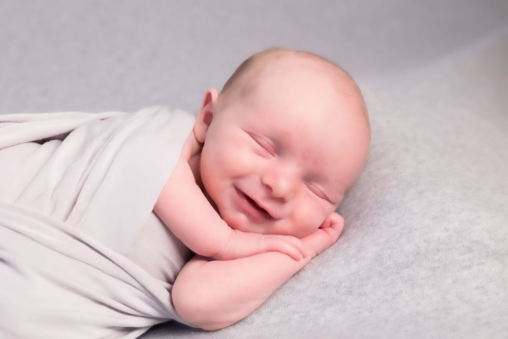 A sleeping baby wrapped in soft gray fabric smiles sweetly while resting on a light colored surface.