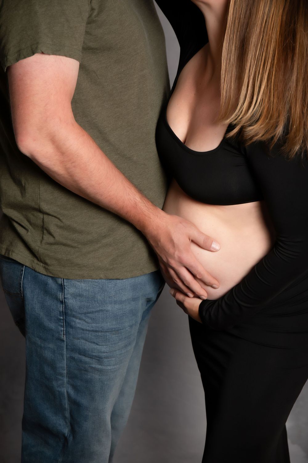 A couple embraces tenderly with hands resting on a pregnant belly, wearing olive green and black clothing against a gray background.