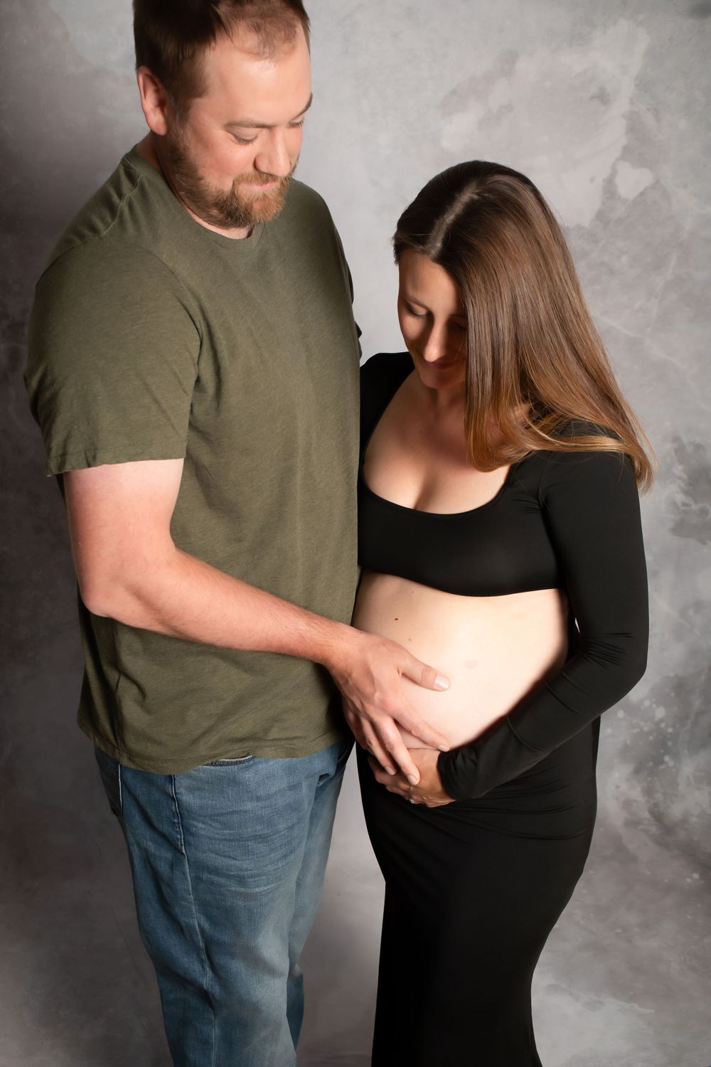 Intimate maternity photo with couple in casual attire against gray backdrop gently touching pregnant belly.