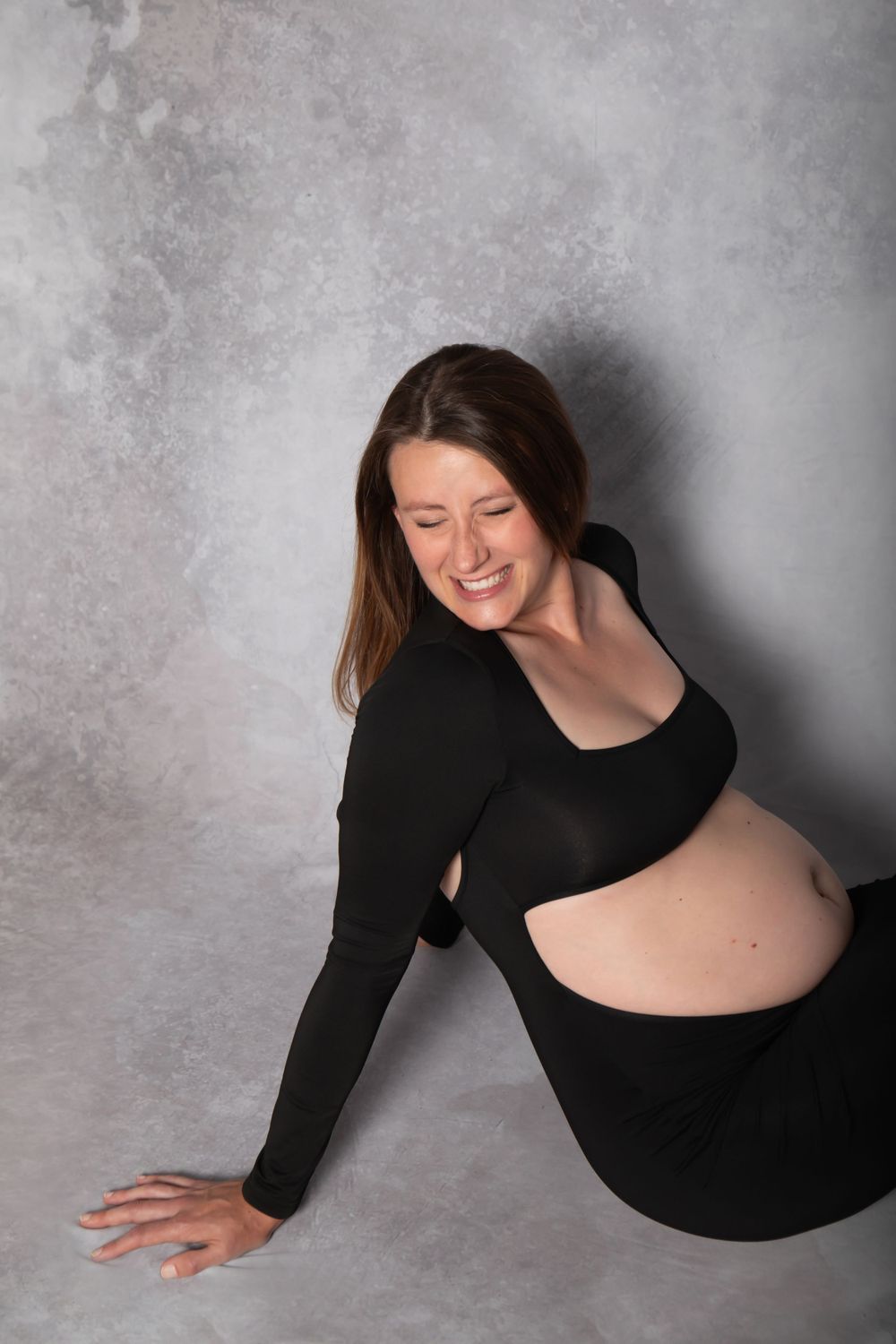A woman in a black crop top and skirt poses against a gray backdrop during a maternity photoshoot.