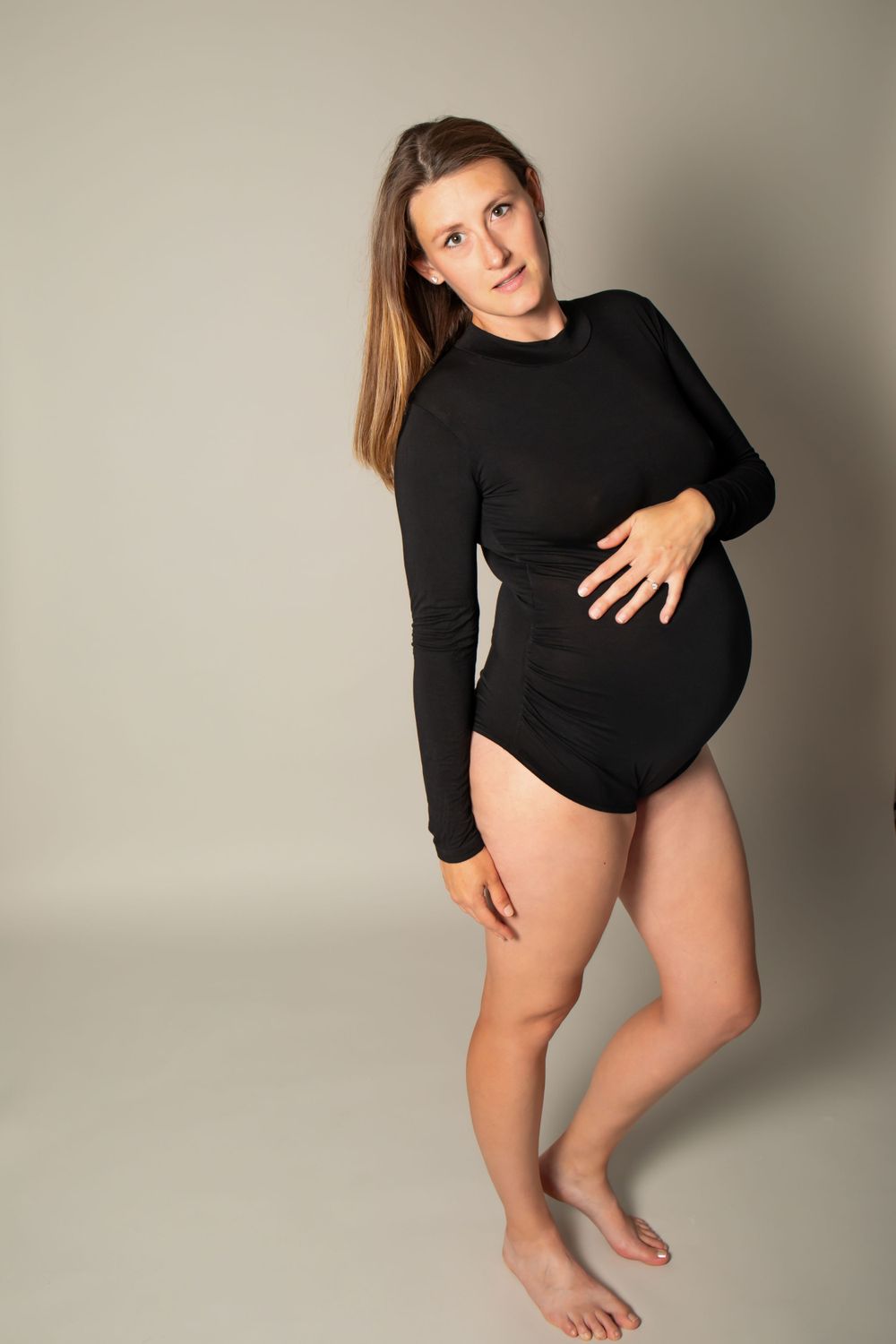 Studio maternity portrait of someone in a black long-sleeve bodysuit posing against a neutral background.