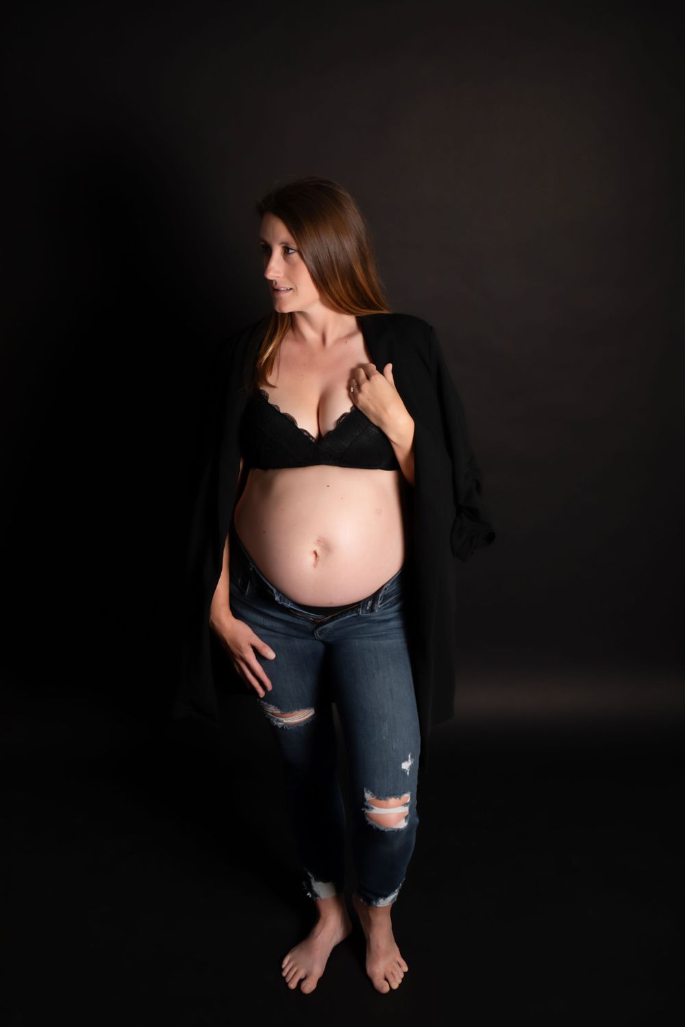 Pregnant woman in distressed jeans and black top poses barefoot against dark backdrop during maternity photoshoot.