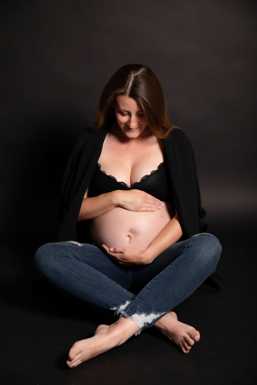 Expectant mother in black top and jeans sits cross-legged on dark background during maternity photoshoot.