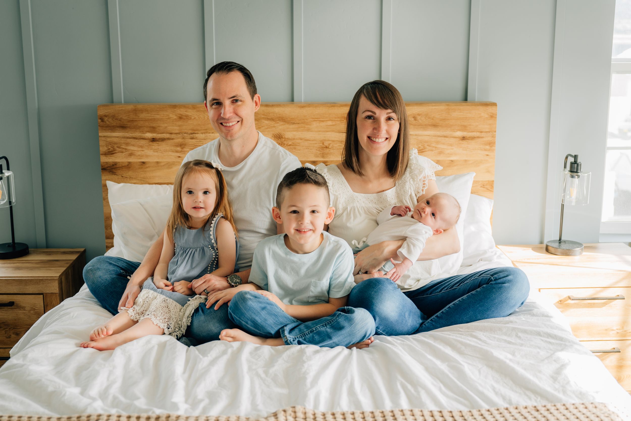Family members sitting together on white bedding with wooden headboard in bright bedroom setting.