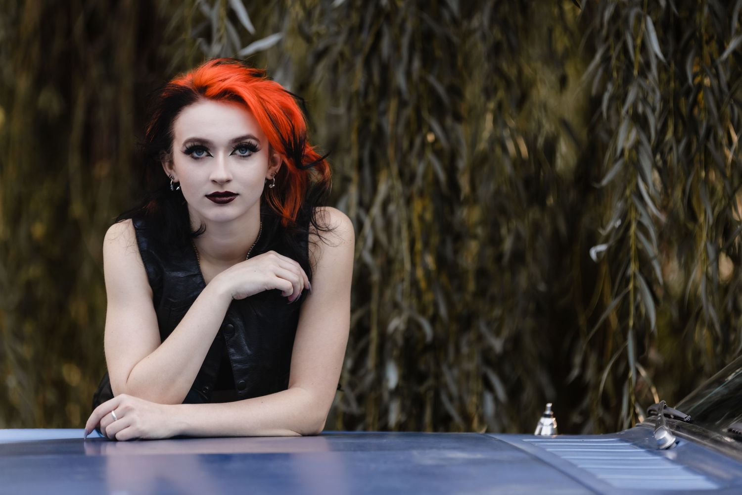 A gothic style portrait with bright orange hair and dark makeup against a willow tree background.