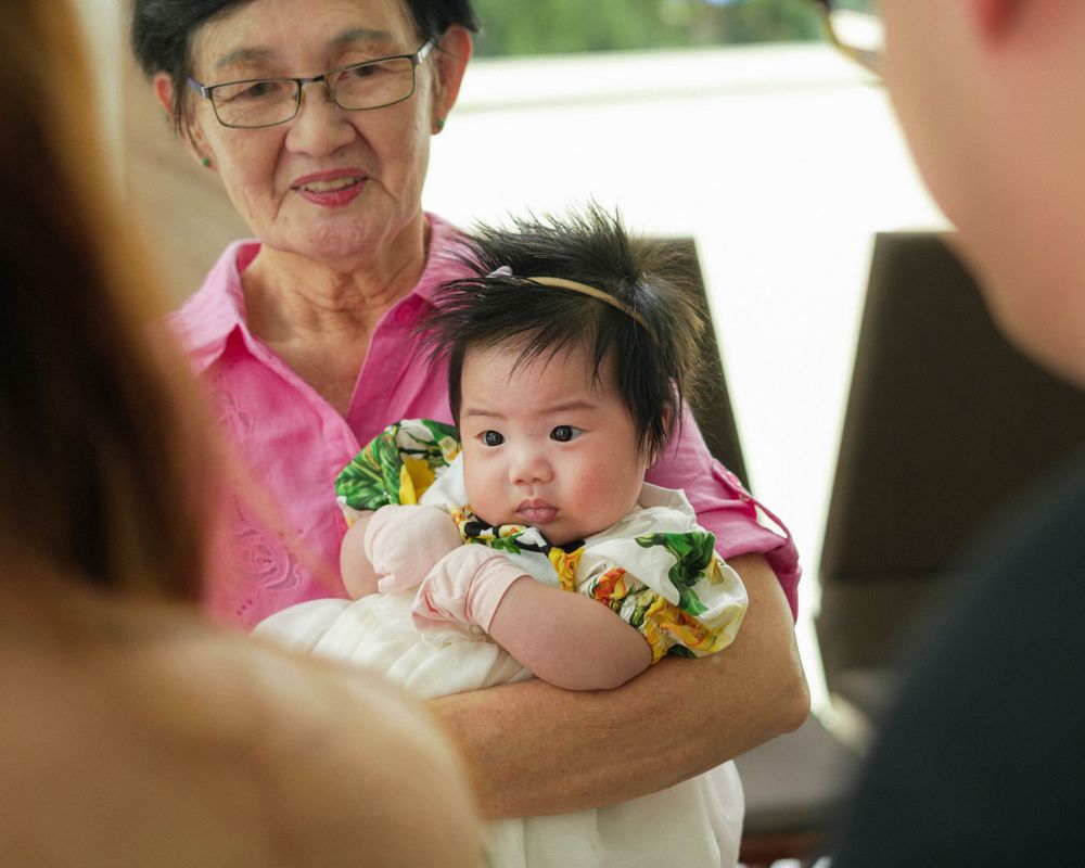 Baby in white dress being held at a family celebration.