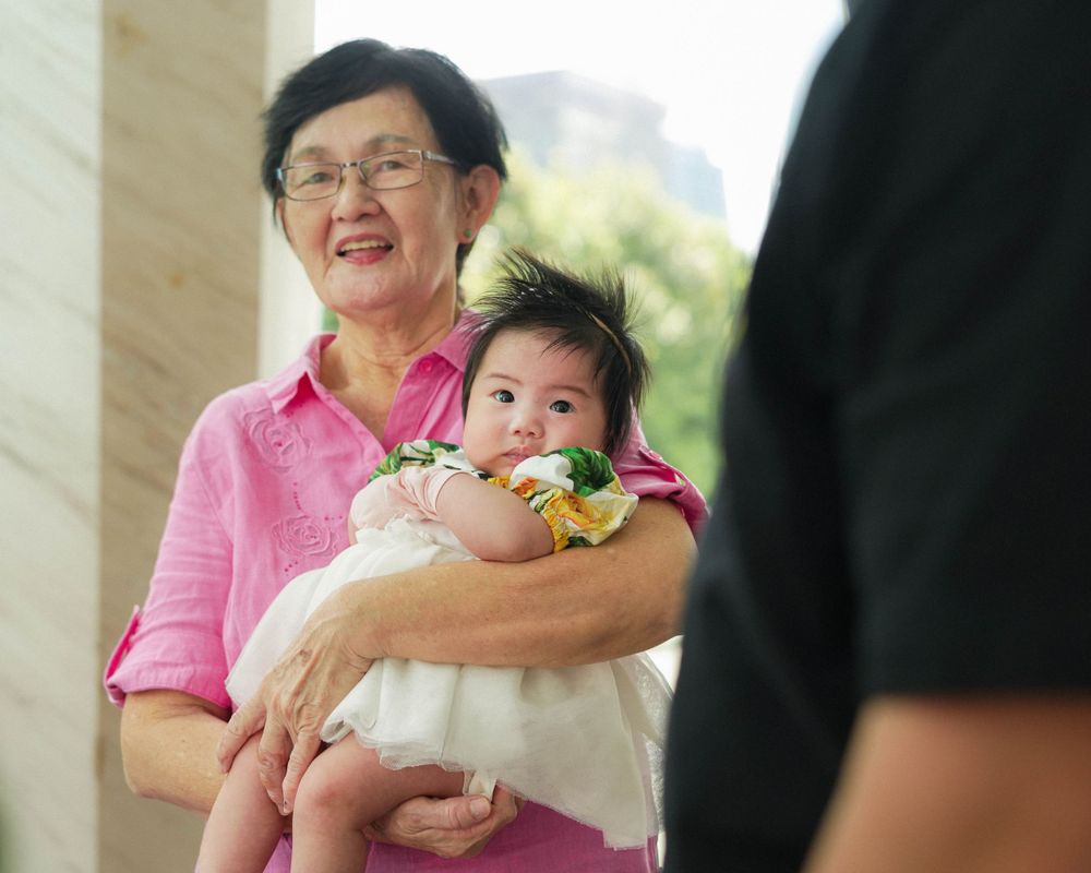 Smiling baby in white dress at a family gathering.