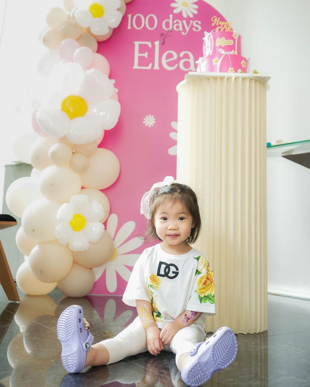 Toddler sitting by pink backdrop with white floral decorations wearing casual attire.