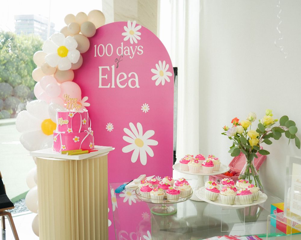 Pink and white themed dessert table with floral decorations and celebration backdrop.