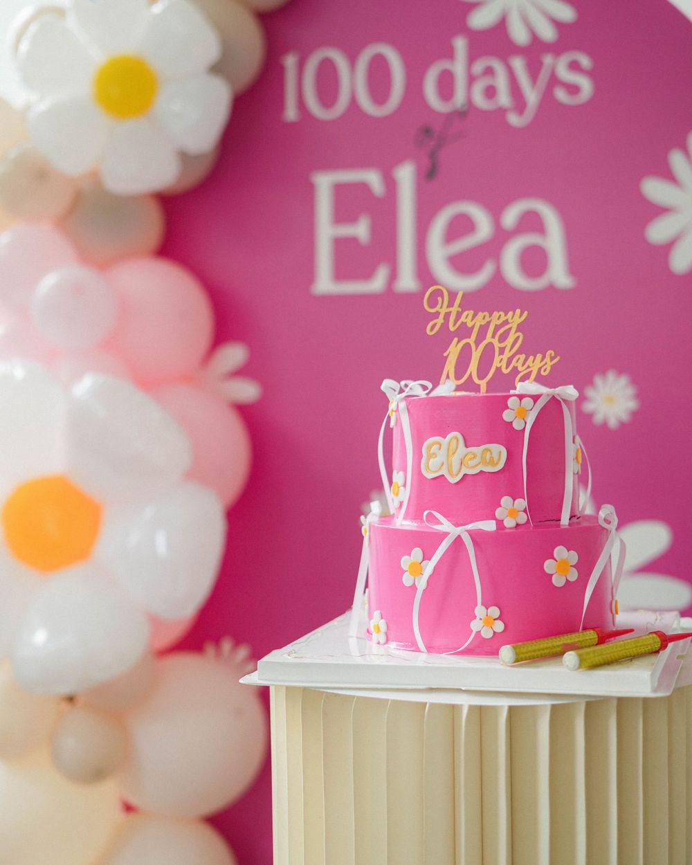 Pink celebration cake with floral designs displayed on pedestal against decorated backdrop.