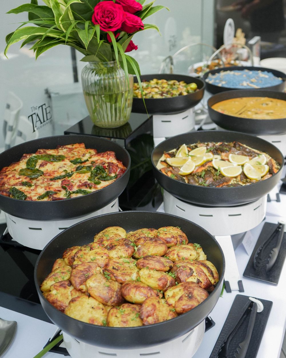 Several hot dishes served in black pans on warming plates with a red rose centerpiece.