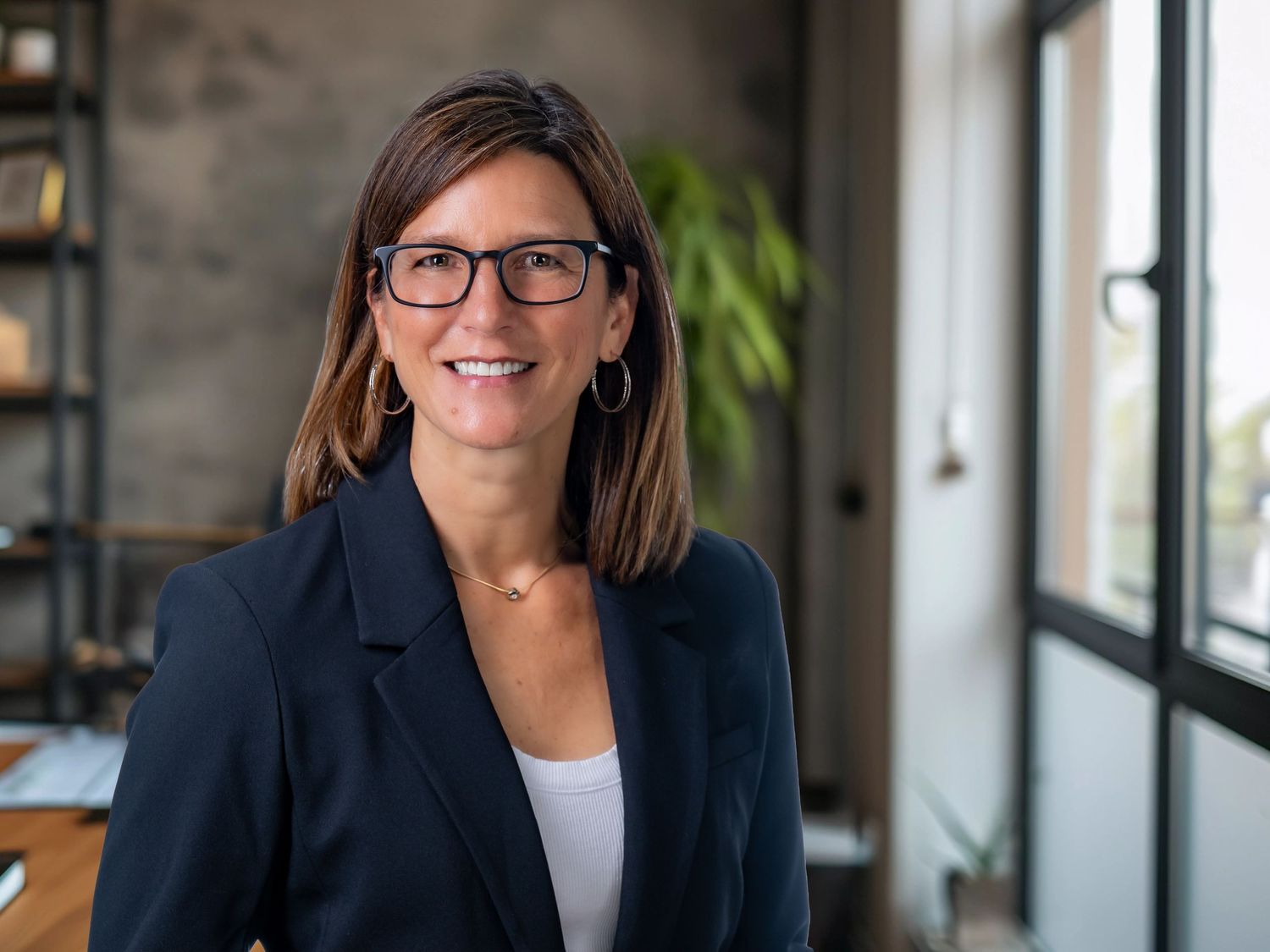 Professional businesswoman in navy blazer and glasses smiling in modern office setting near window.