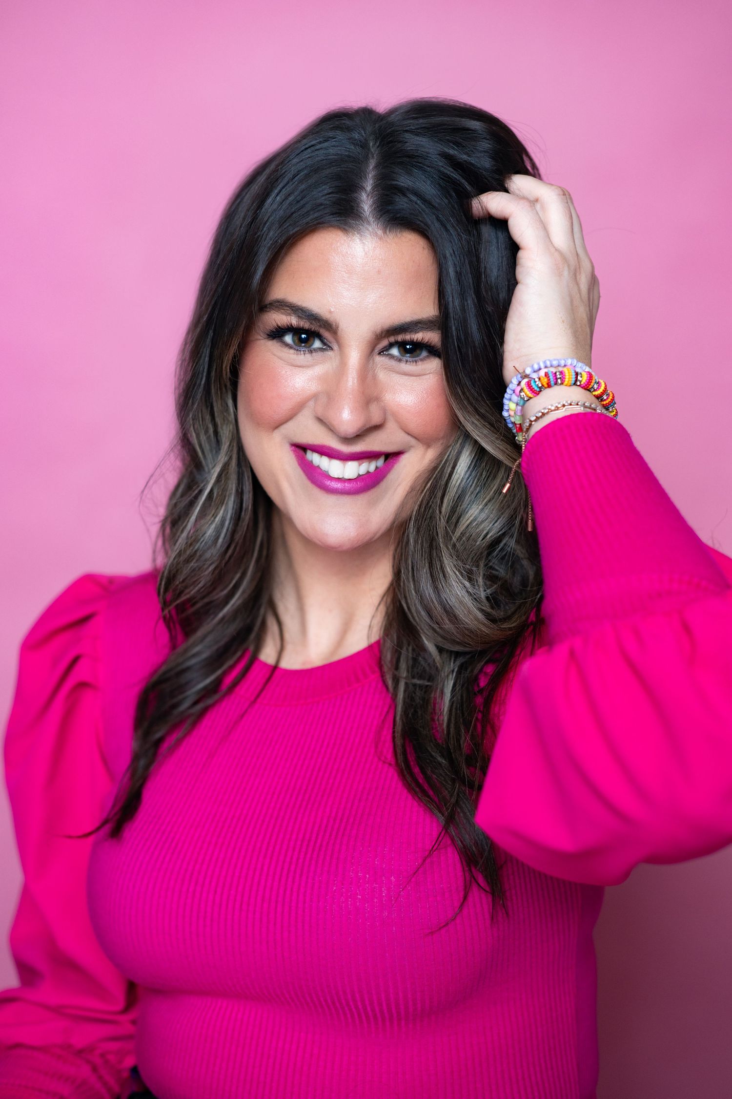 Someone with long dark wavy hair poses in a bright pink sweater against a pink background.