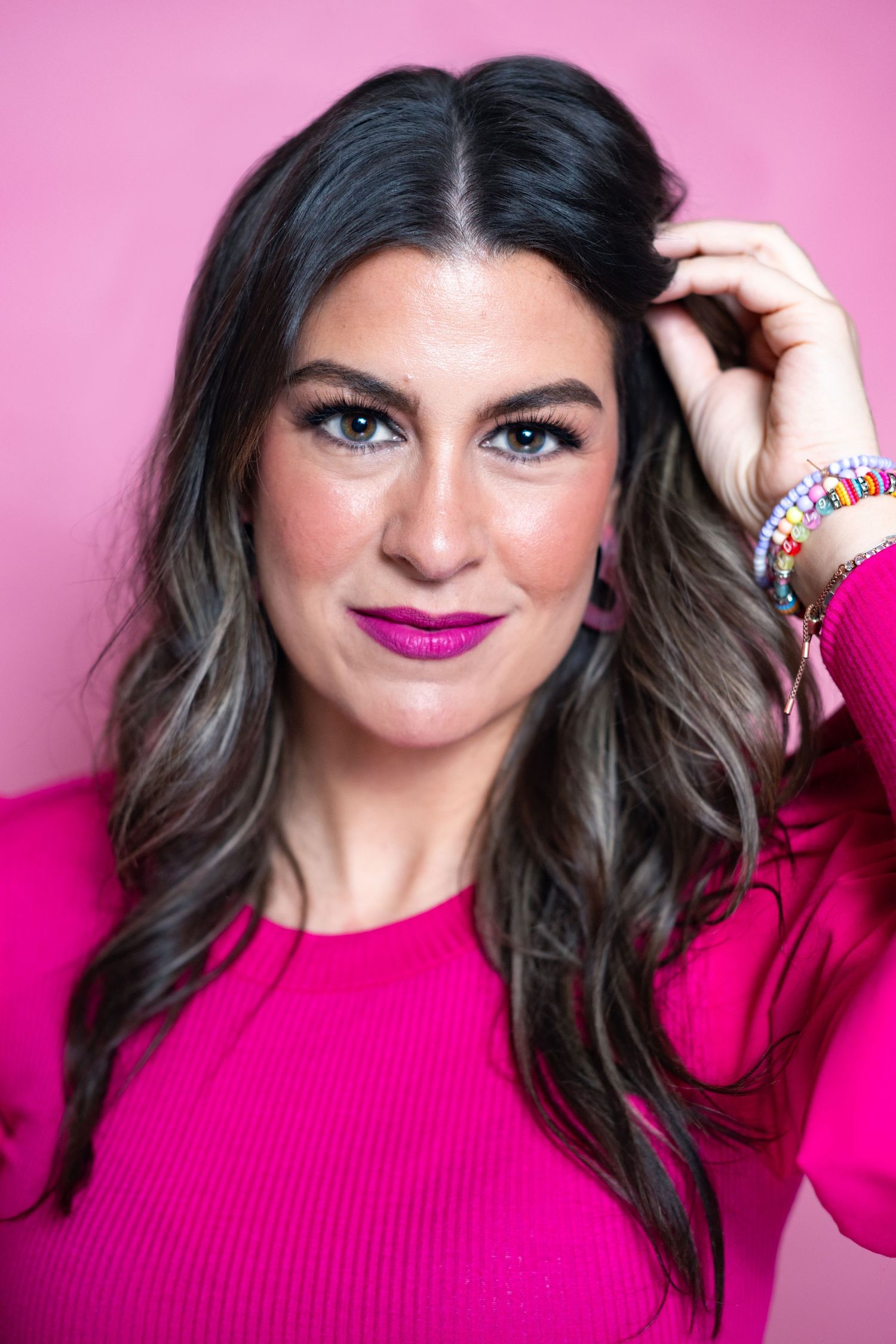 Portrait against pink background showing dark wavy hair, bright magenta lipstick, and matching fuchsia sweater.