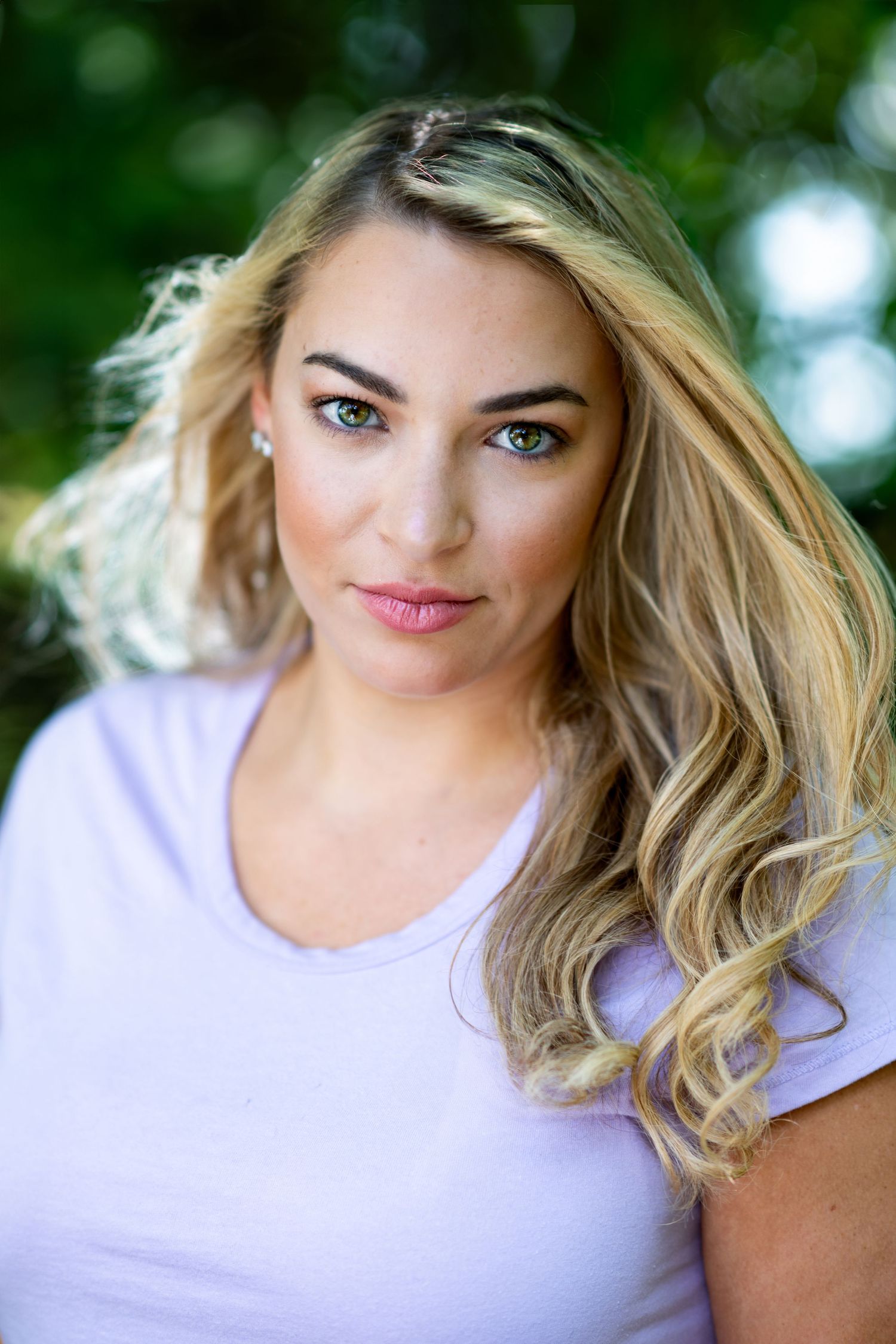 Outdoor portrait with soft natural lighting shows blonde wavy hair and a light purple t-shirt against a blurred green background.