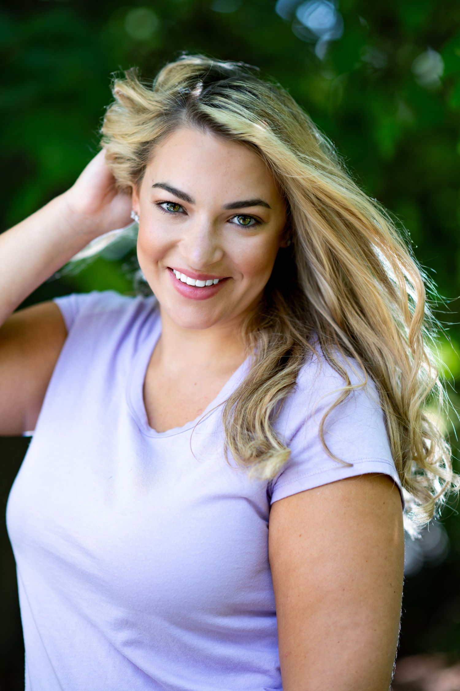 A smiling blonde woman in a light purple t-shirt poses outdoors with windswept hair against a natural green background.