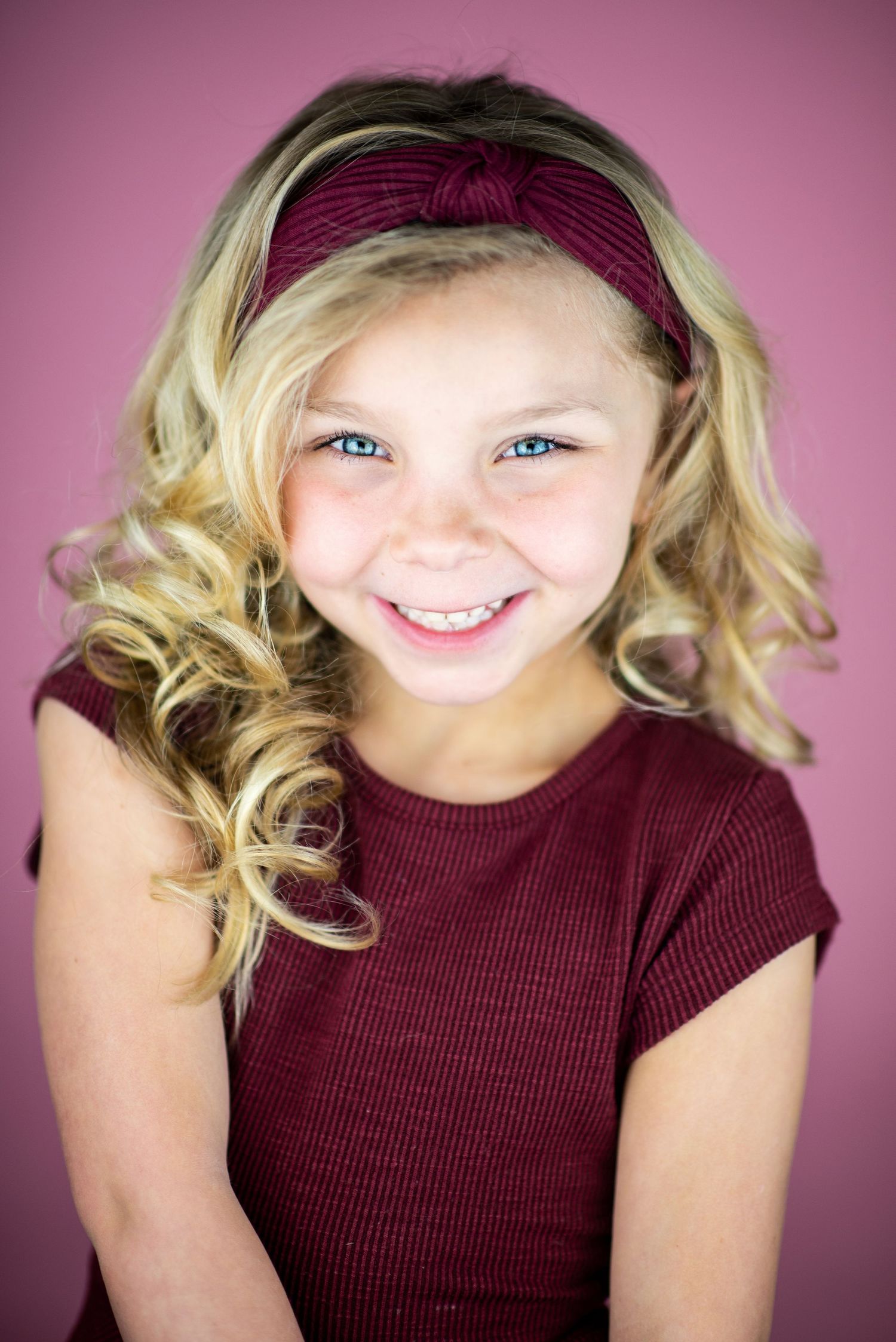 Portrait of a young blonde person in a burgundy top and matching headband smiling warmly against a pink backdrop.