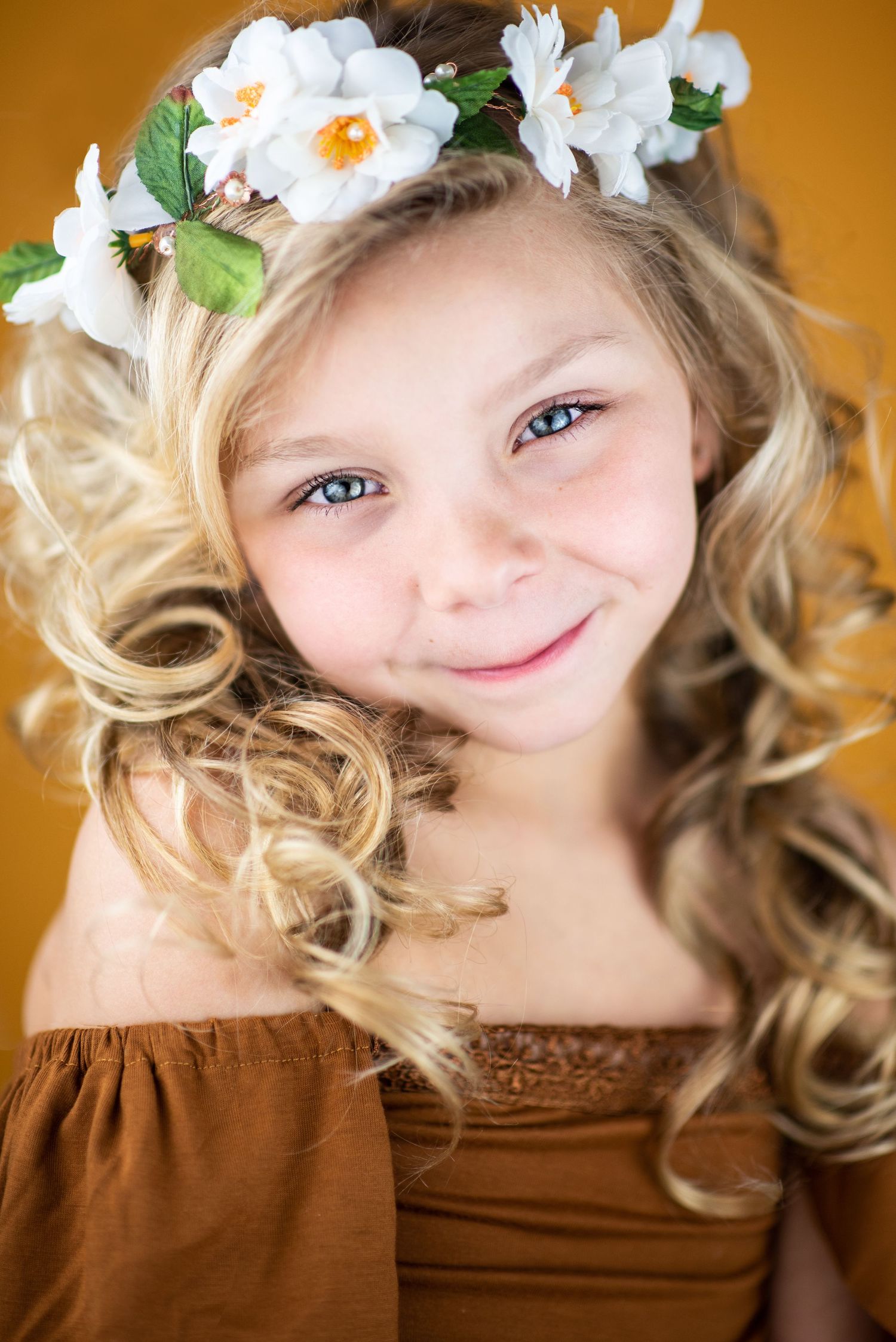 Portrait with blonde curls wearing a brown dress and white flower crown crown against an orange background.