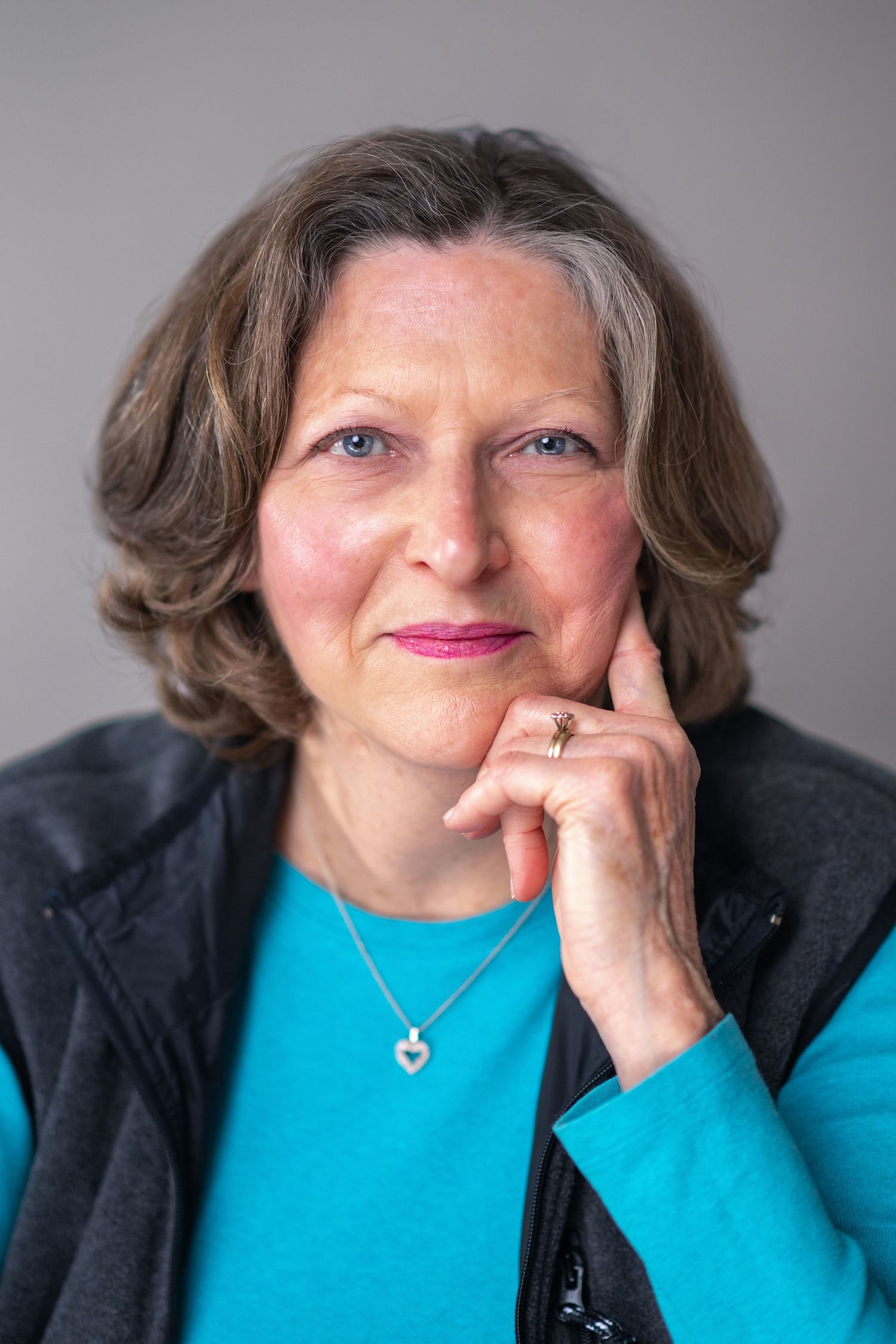 Portrait of someone in a teal sweater and dark cardigan posing with hand on chin against gray background.