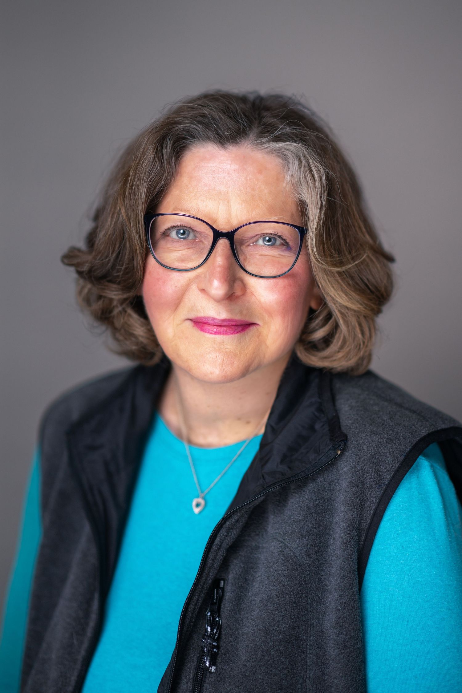 Professional portrait of a person wearing glasses and a black vest over a turquoise sweater against a gray background.