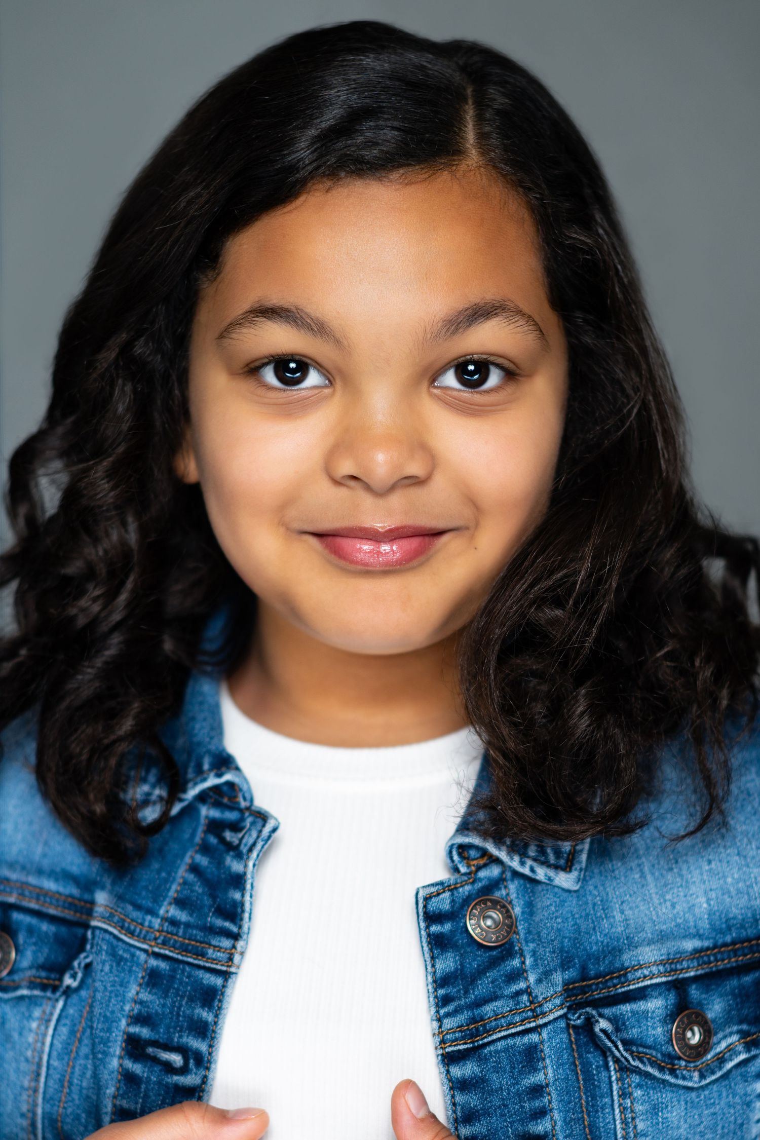Young person wearing a denim jacket and white shirt smiles warmly in a professional headshot photo.
