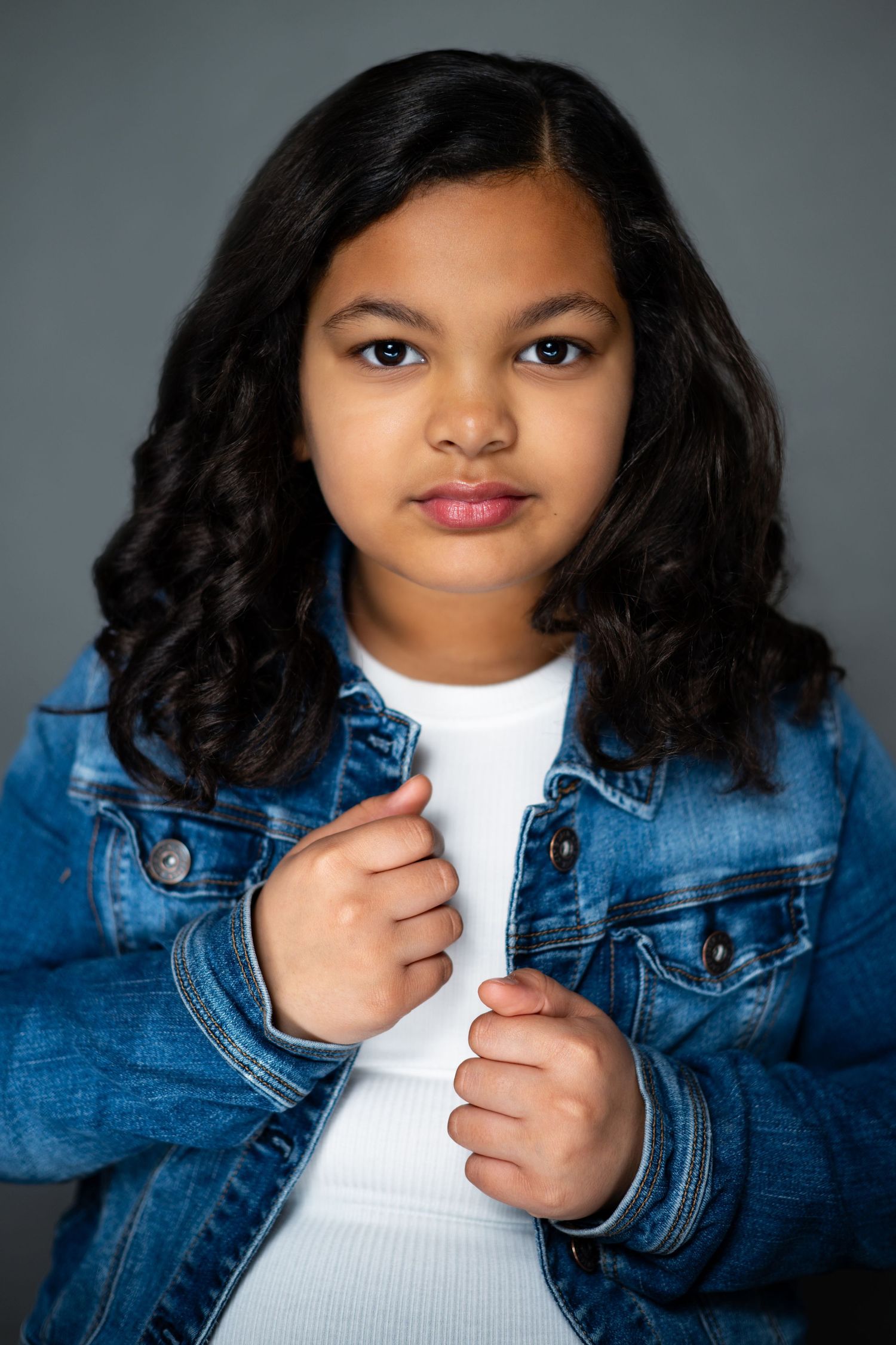 Young person with long dark wavy hair wearing a denim jacket and white shirt against a gray background.