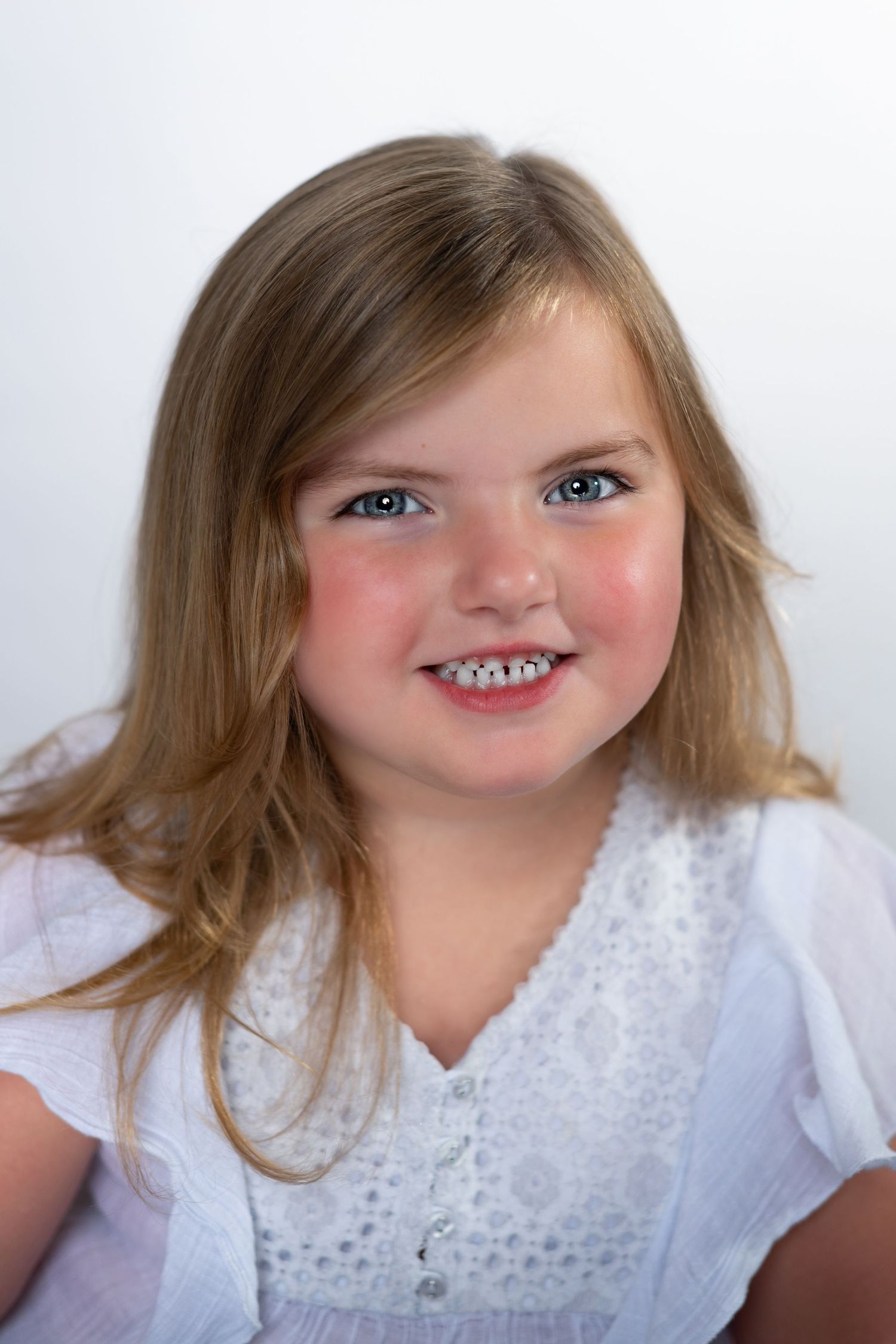Sweet portrait of a child with honey blonde hair wearing a white eyelet lace top against a neutral background.