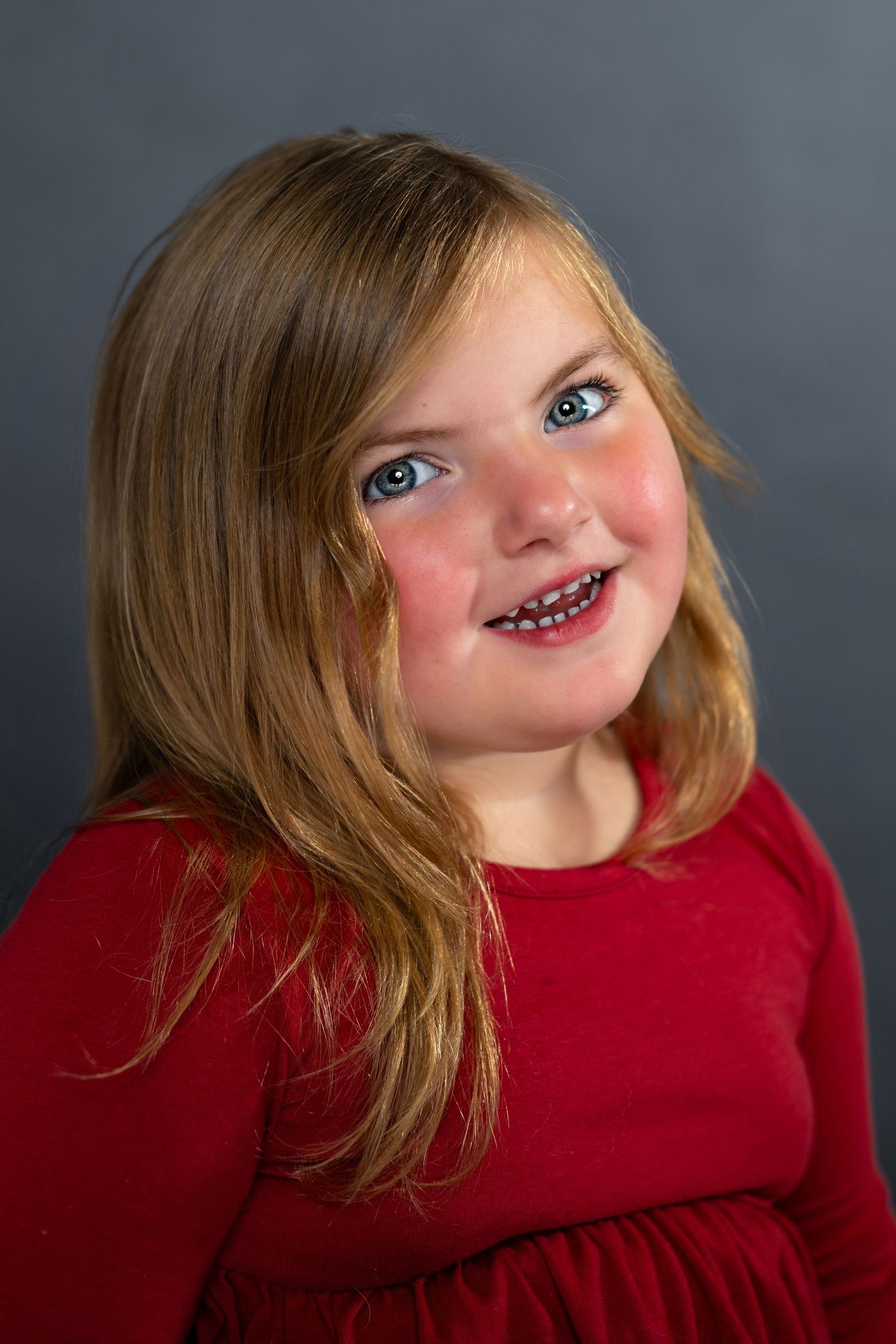 A child in a red dress smiles brightly with blonde hair against a gray studio backdrop.