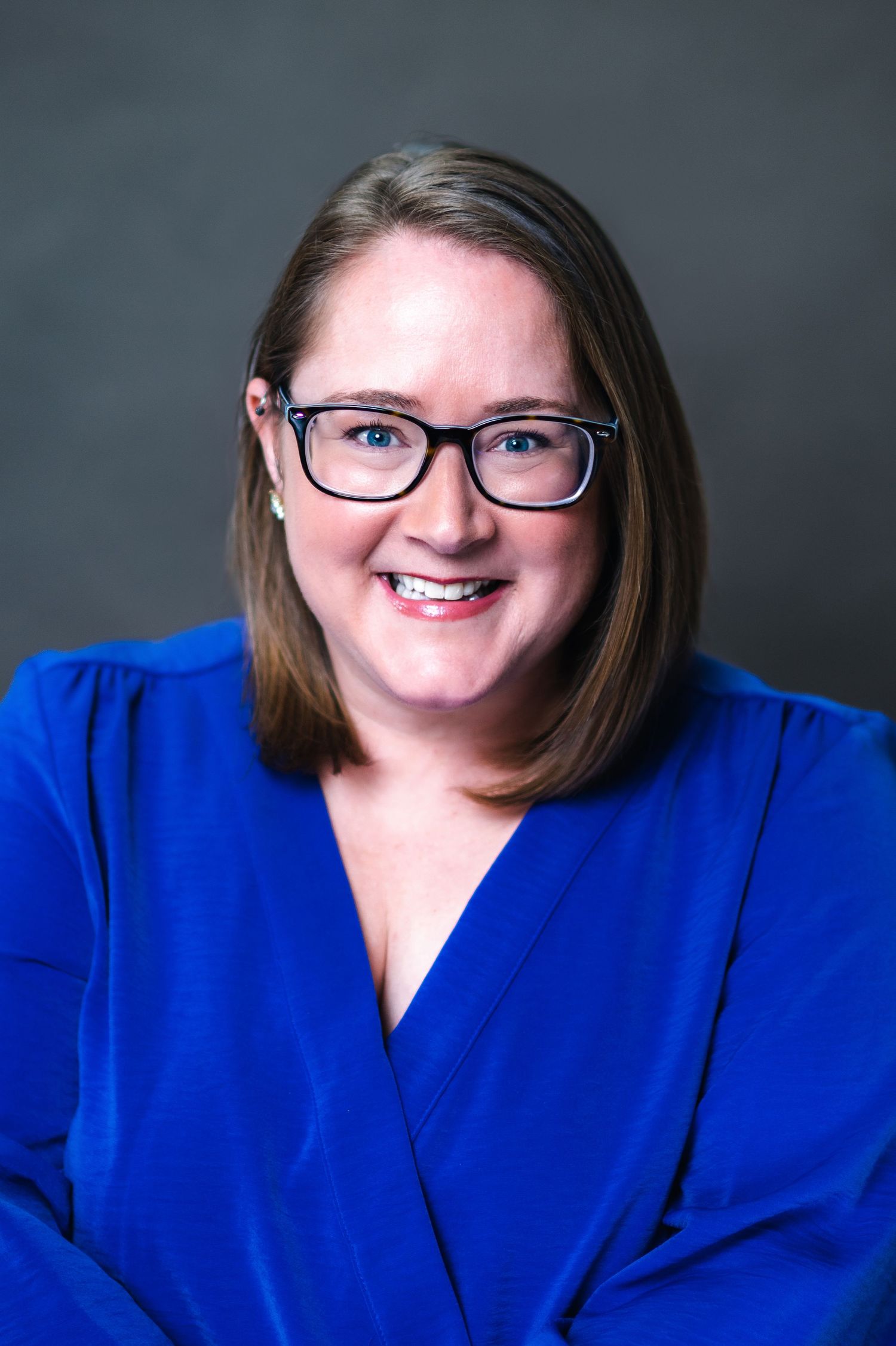 Professional headshot portrait of someone in a bright royal blue blazer and glasses against a dark background.