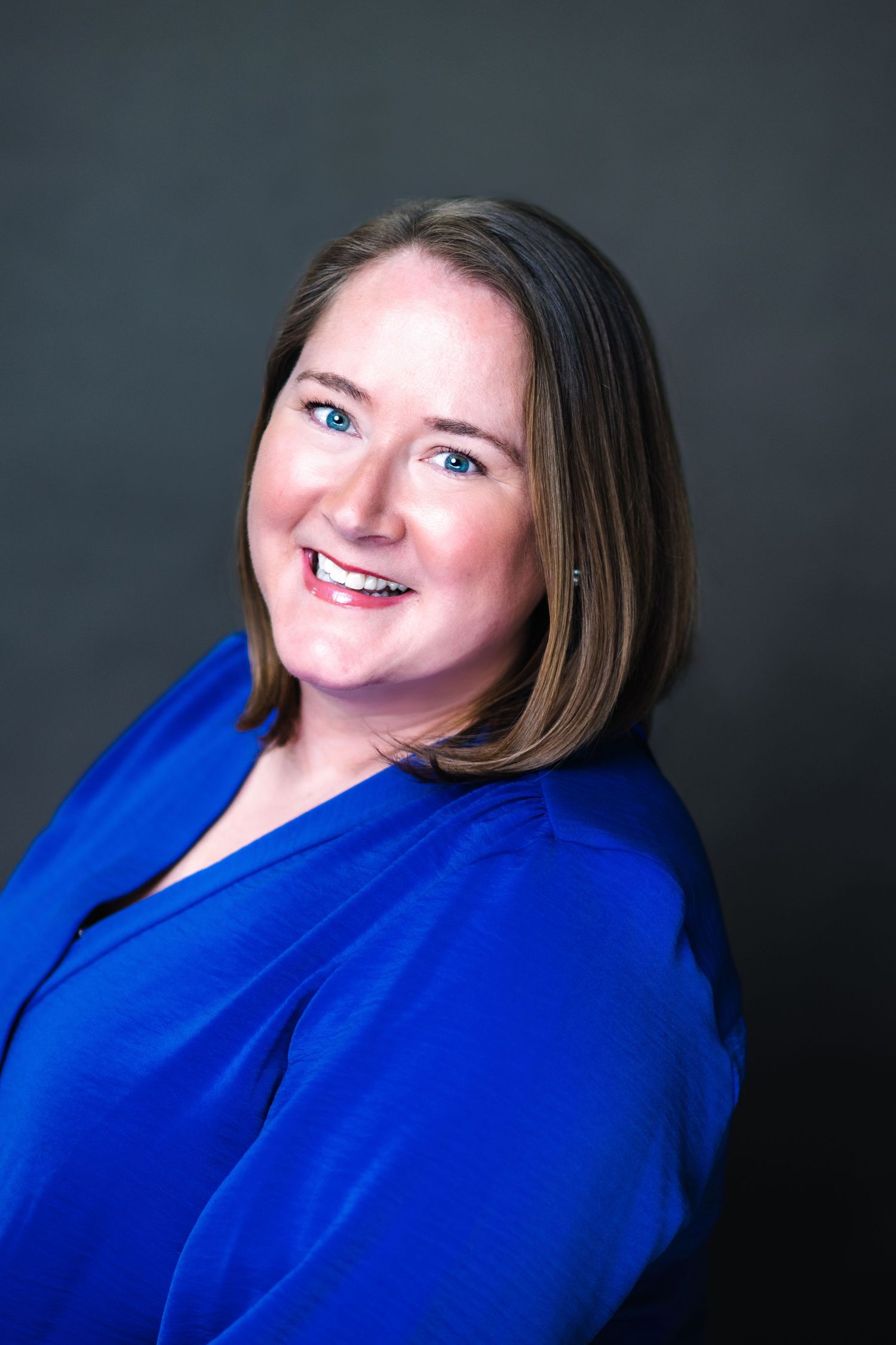 Professional headshot portrait of someone wearing a bright royal blue blazer against a dark gray background with a warm smile.