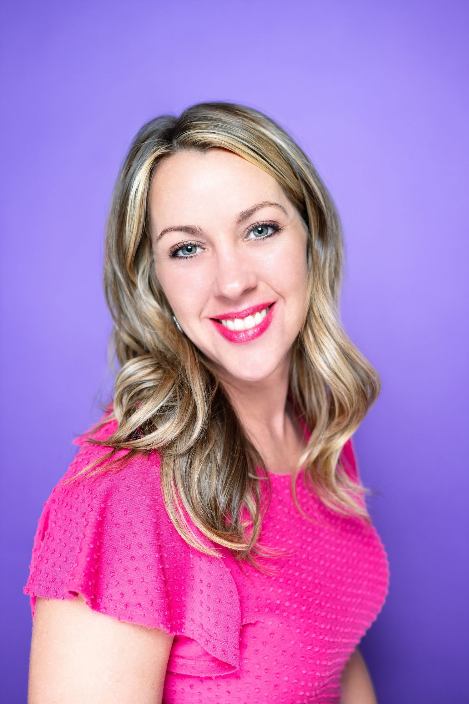 Professional headshot of someone wearing a bright pink top against a purple background, smiling warmly at the camera.