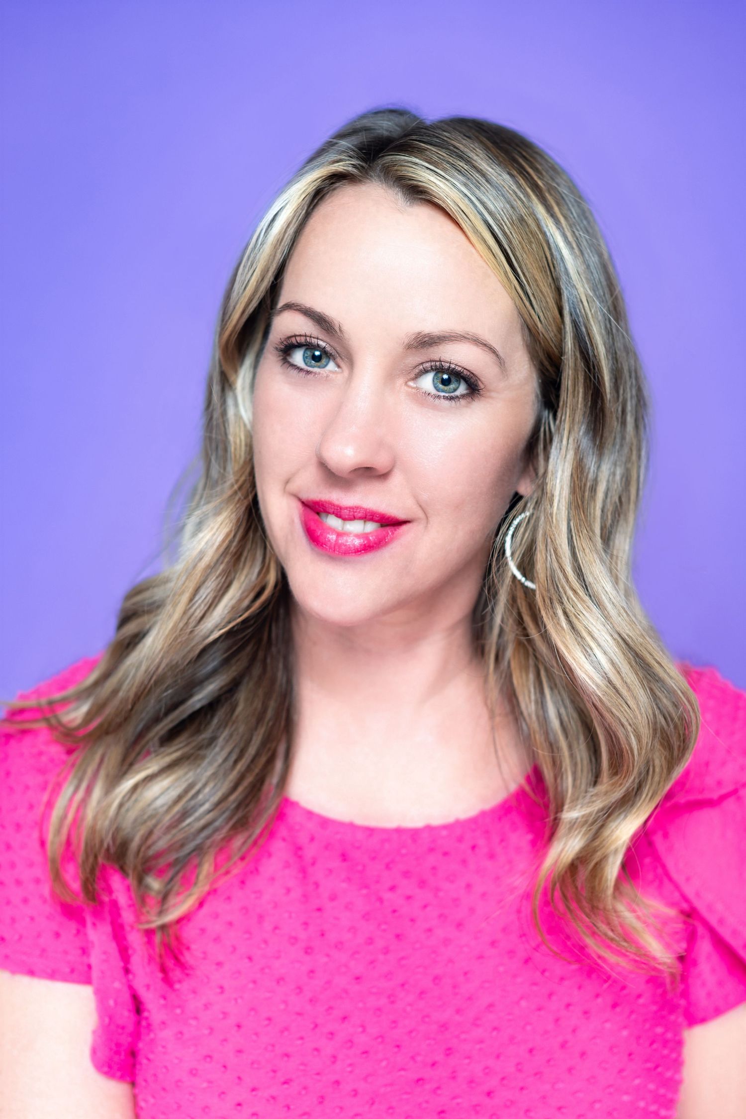 Professional headshot with blonde wavy hair wearing bright pink top against purple background.