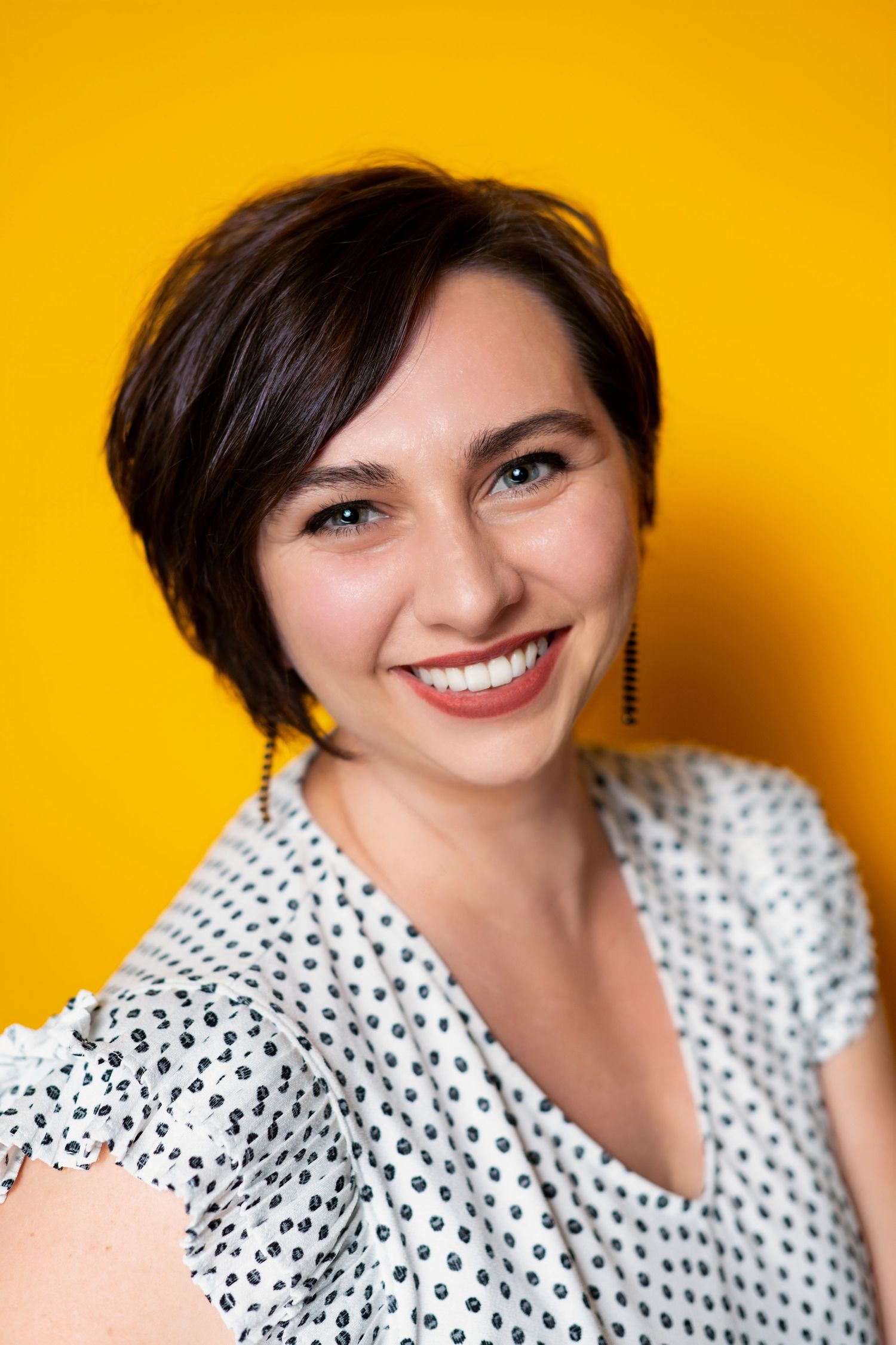 Portrait against vibrant yellow background showing warm smile and short dark hair, wearing polka dot top.