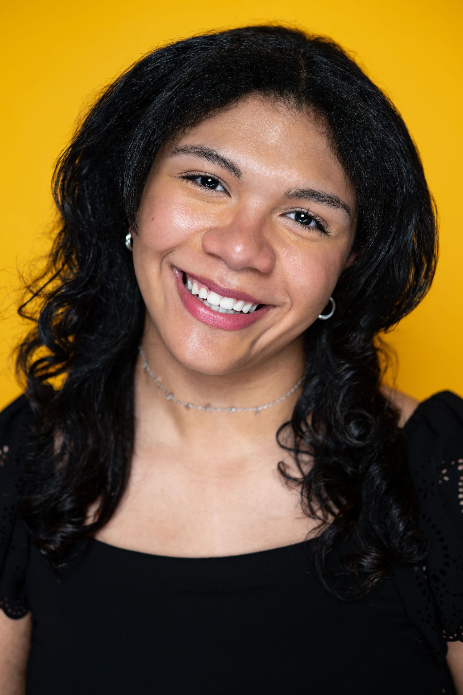Portrait of someone with long dark curly hair wearing a black top and smiling warmly against a bright yellow background.