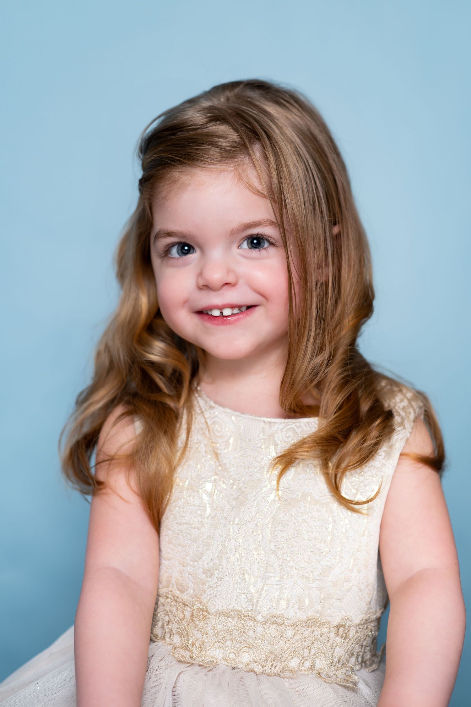 A sweet portrait of a young child wearing a cream-colored dress with lace details against a light blue background.