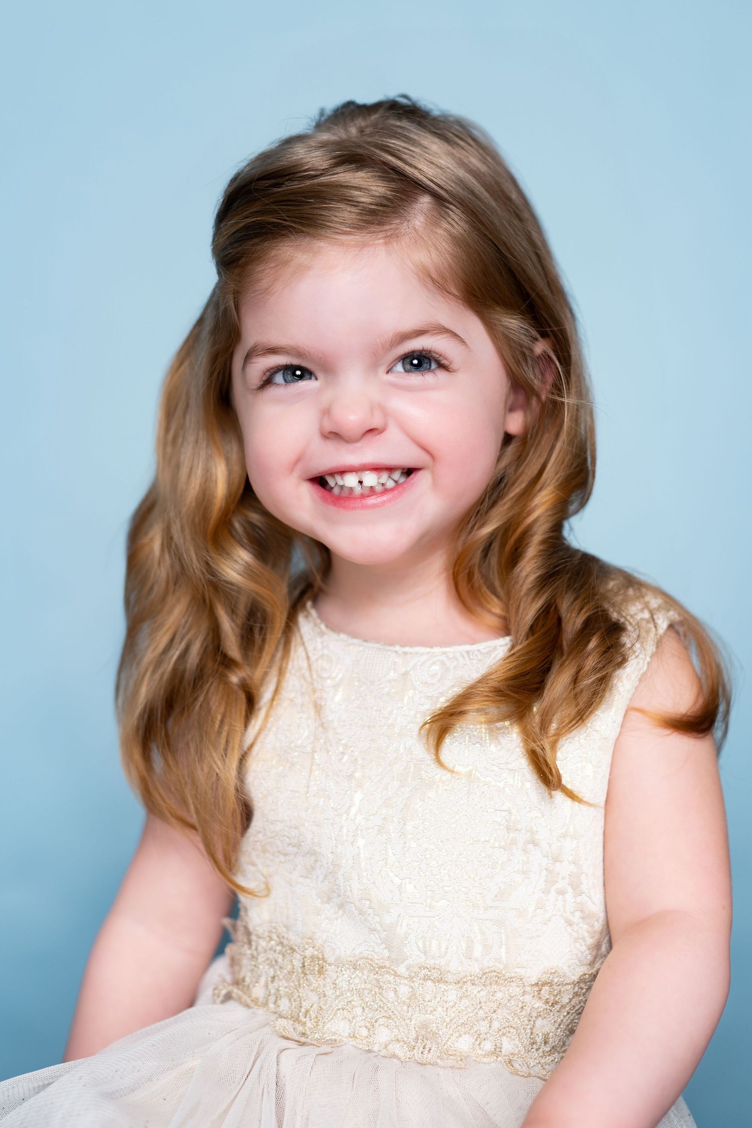 Young child wearing a cream-colored dress with lace detailing smiles brightly against a light blue backdrop.
