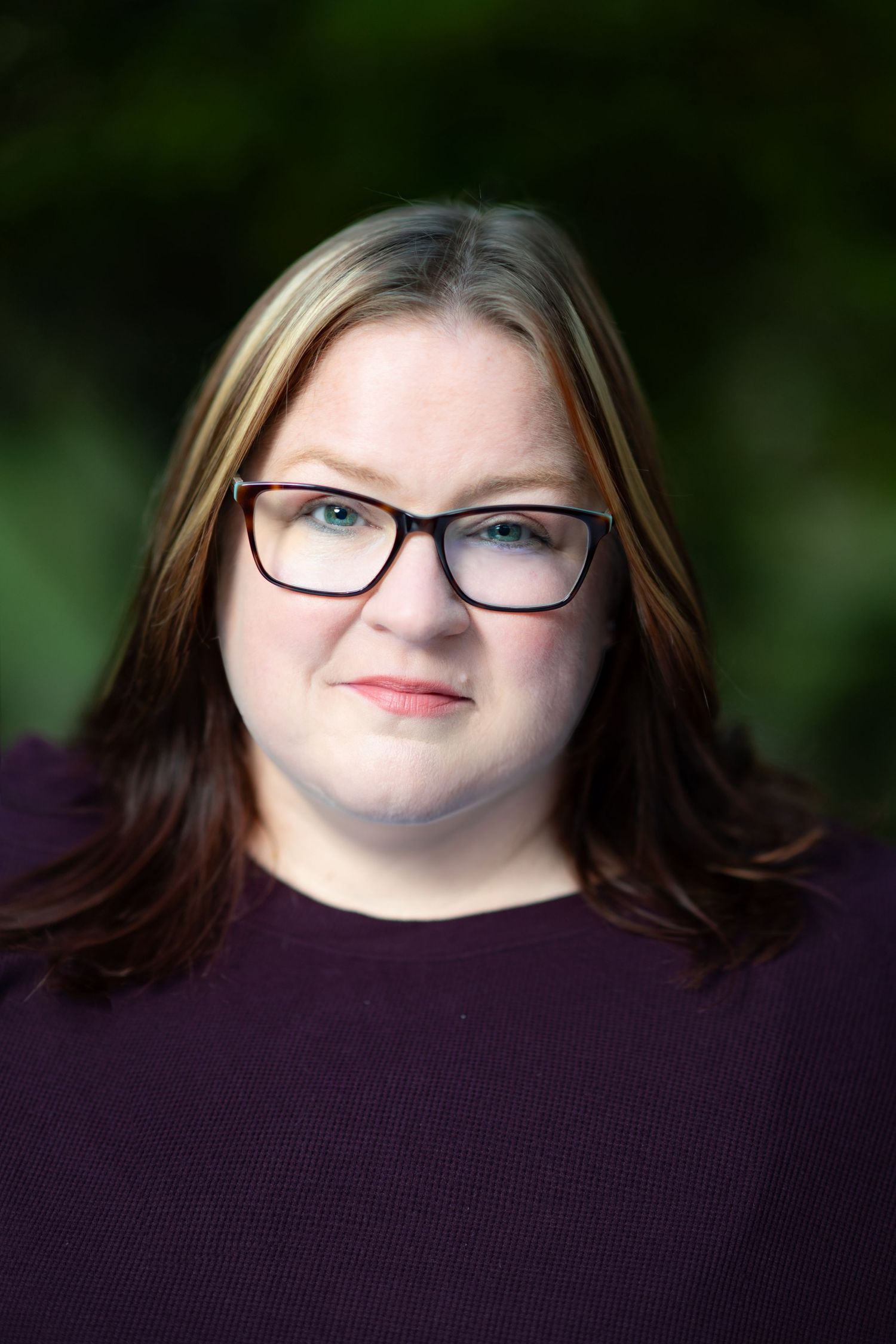 Portrait of someone wearing black frame glasses and a purple sweater against a blurred green background.