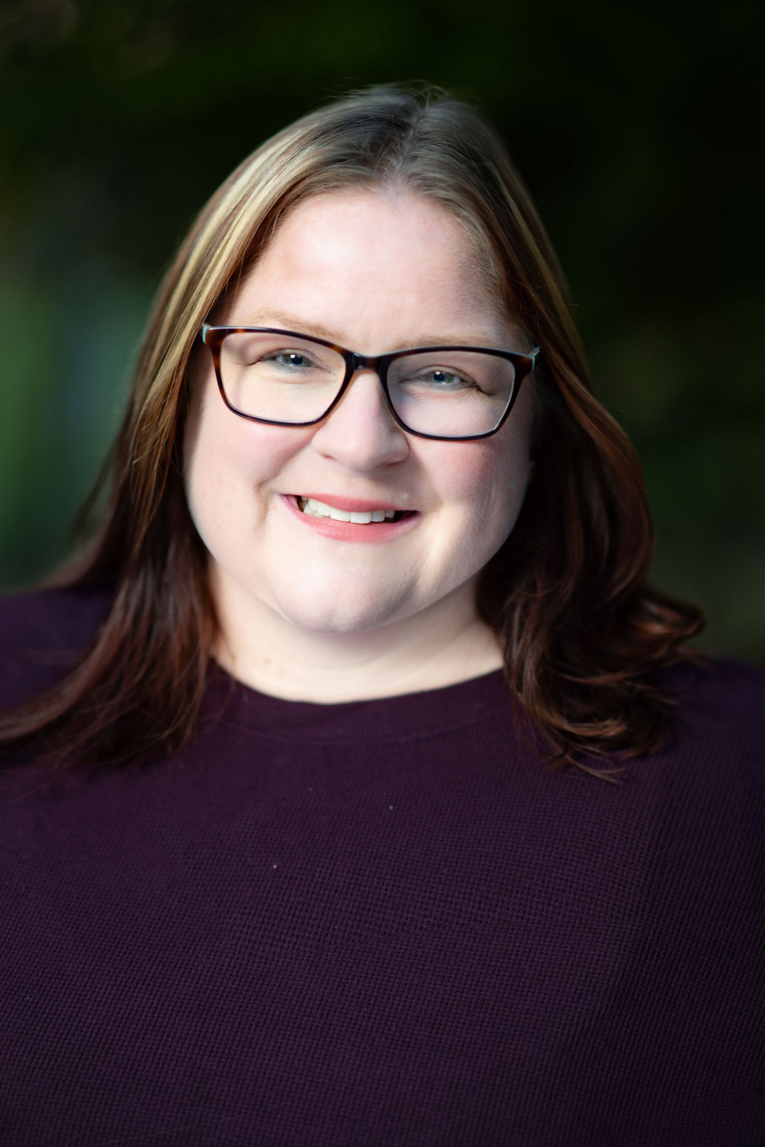 Professional headshot portrait of someone wearing glasses and a purple sweater with a soft-focus natural background.