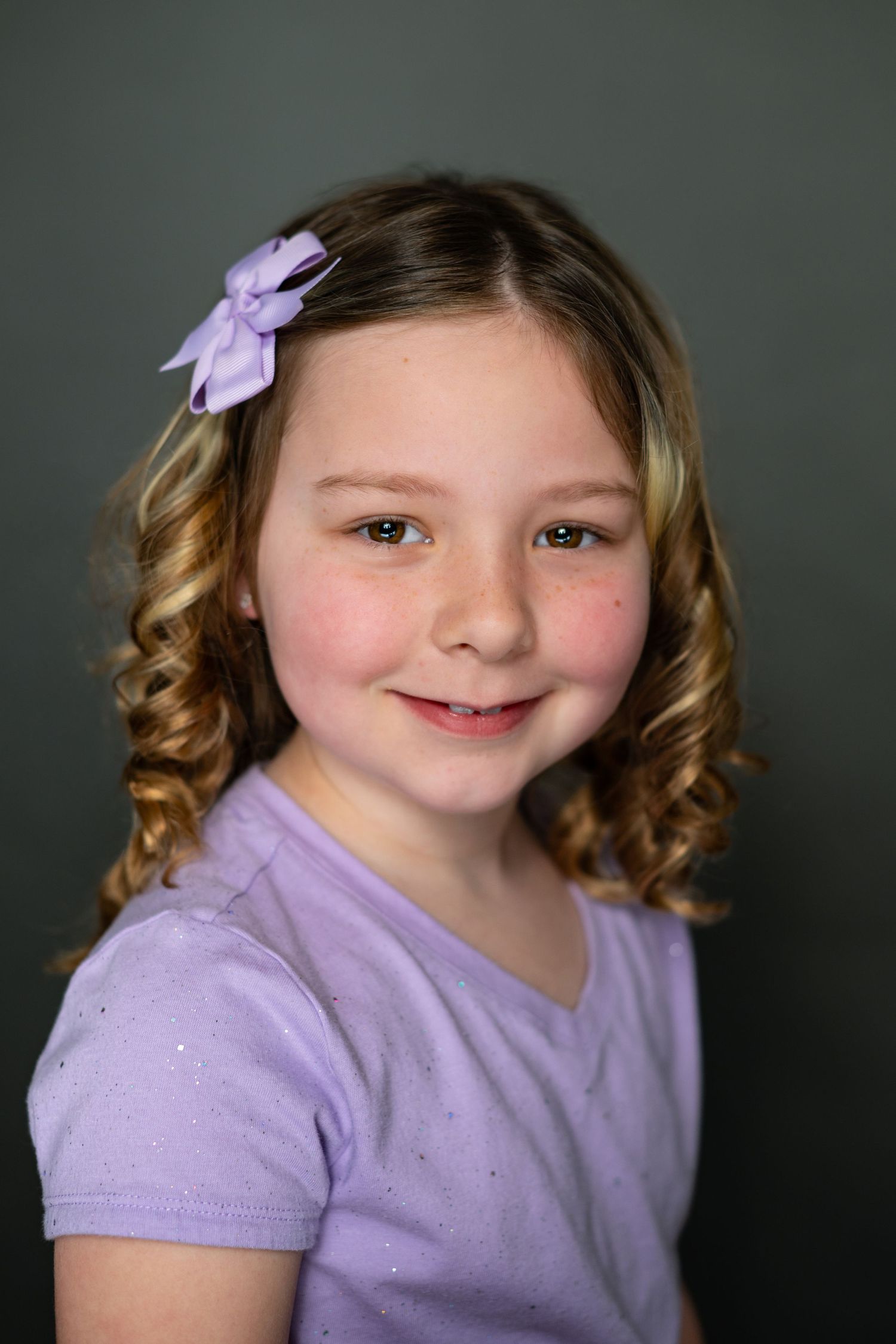 Portrait of a young child in a purple shirt with curly hair and a matching lilac bow against a dark background.