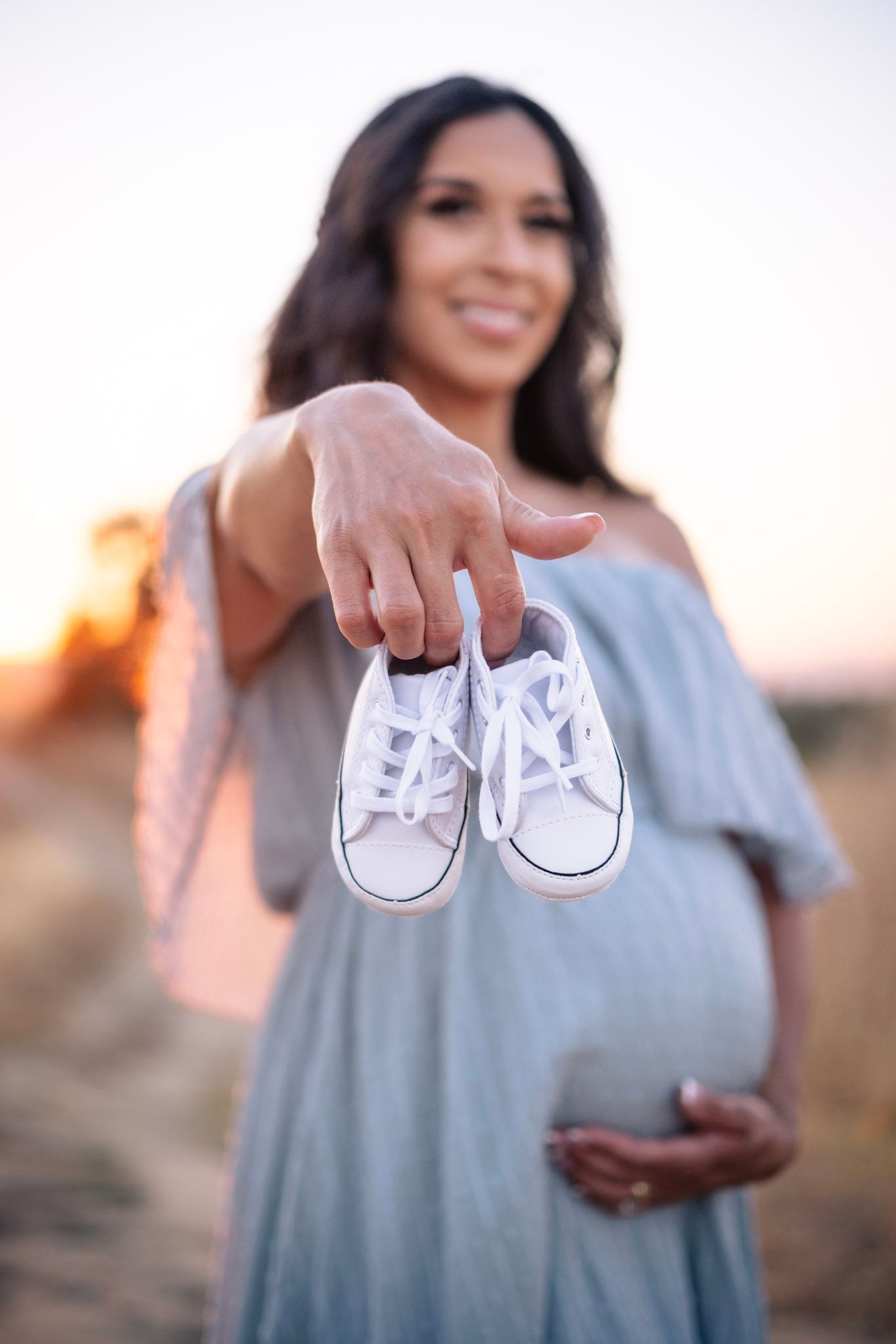 Expectant mother in blue dress holds tiny white baby shoes at sunset while cradling her baby bump.